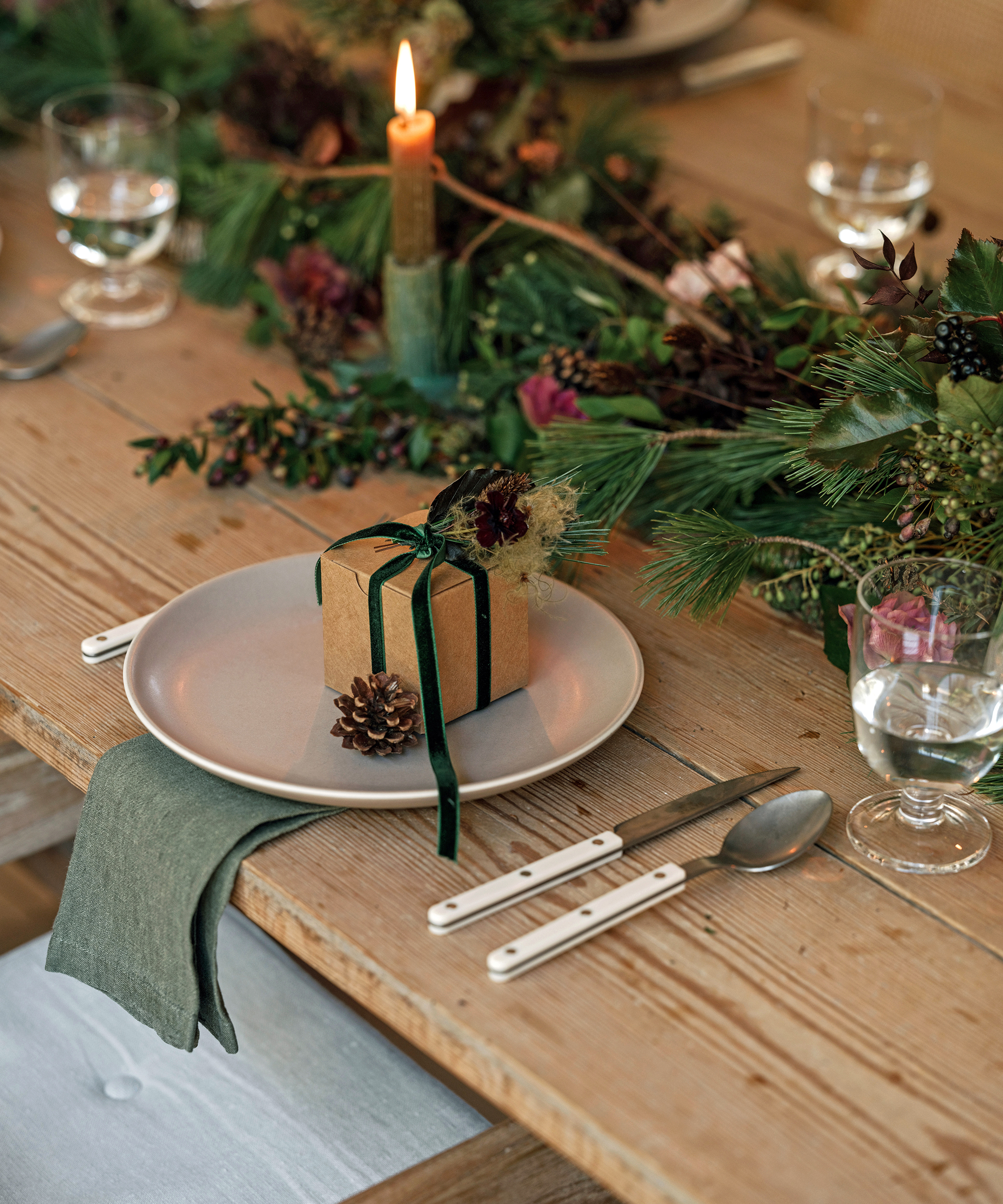 A rustic wooden table set for a vegetarian meal, featuring a plate with a small gift box tied with green ribbon and a pinecone, a green napkin, cutlery, glasses of water, and a festive greenery centerpiece with a lit candle.