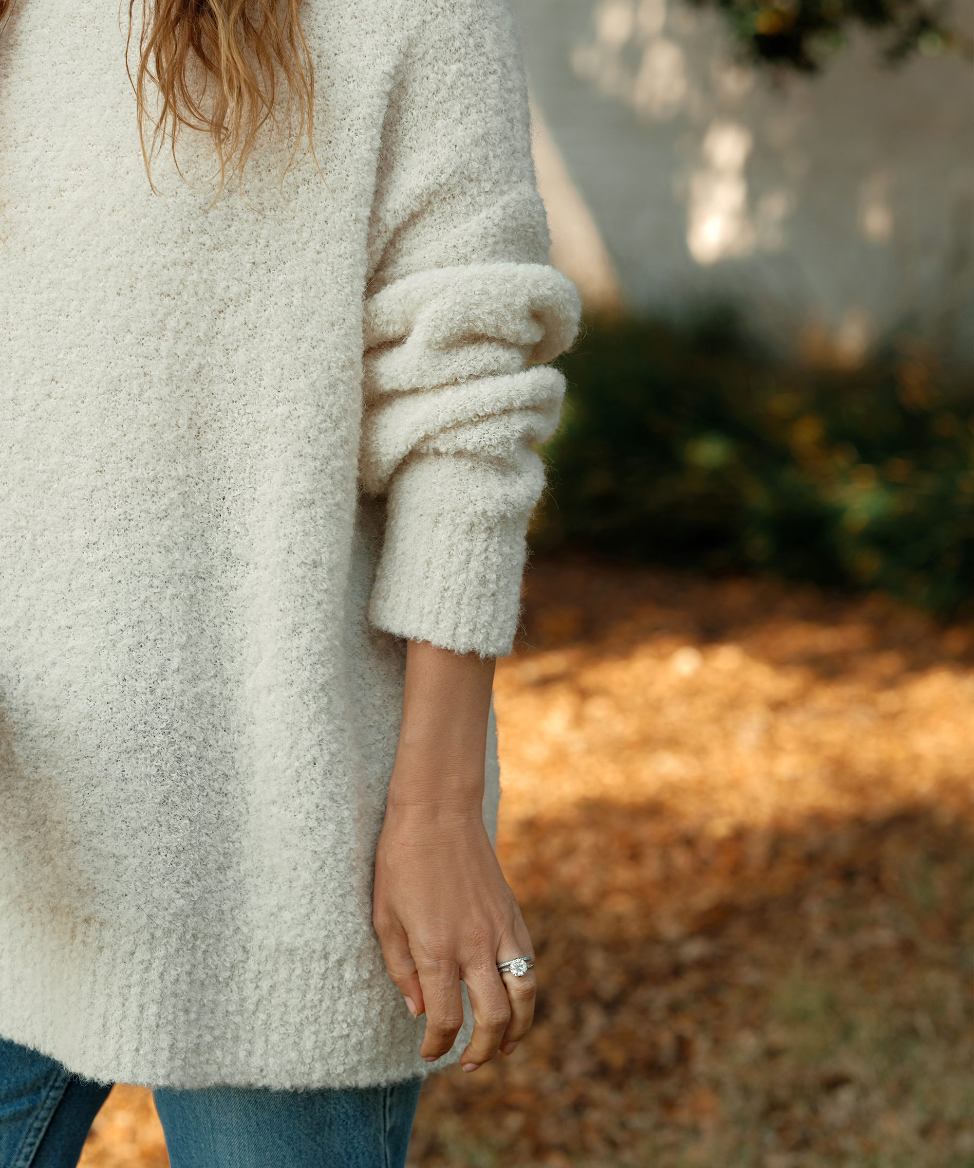 A person wearing a soft, textured cream sweater—an ideal piece for a capsule wardrobe—stands outdoors on a grassy area with dry leaves. Only their torso and one hand with a ring are visible, sleeves partially rolled up.