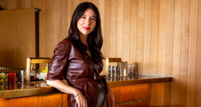 sasha in a maroon leather jumpsuit leaning against a wood bar with leather stools in a wood paneled room