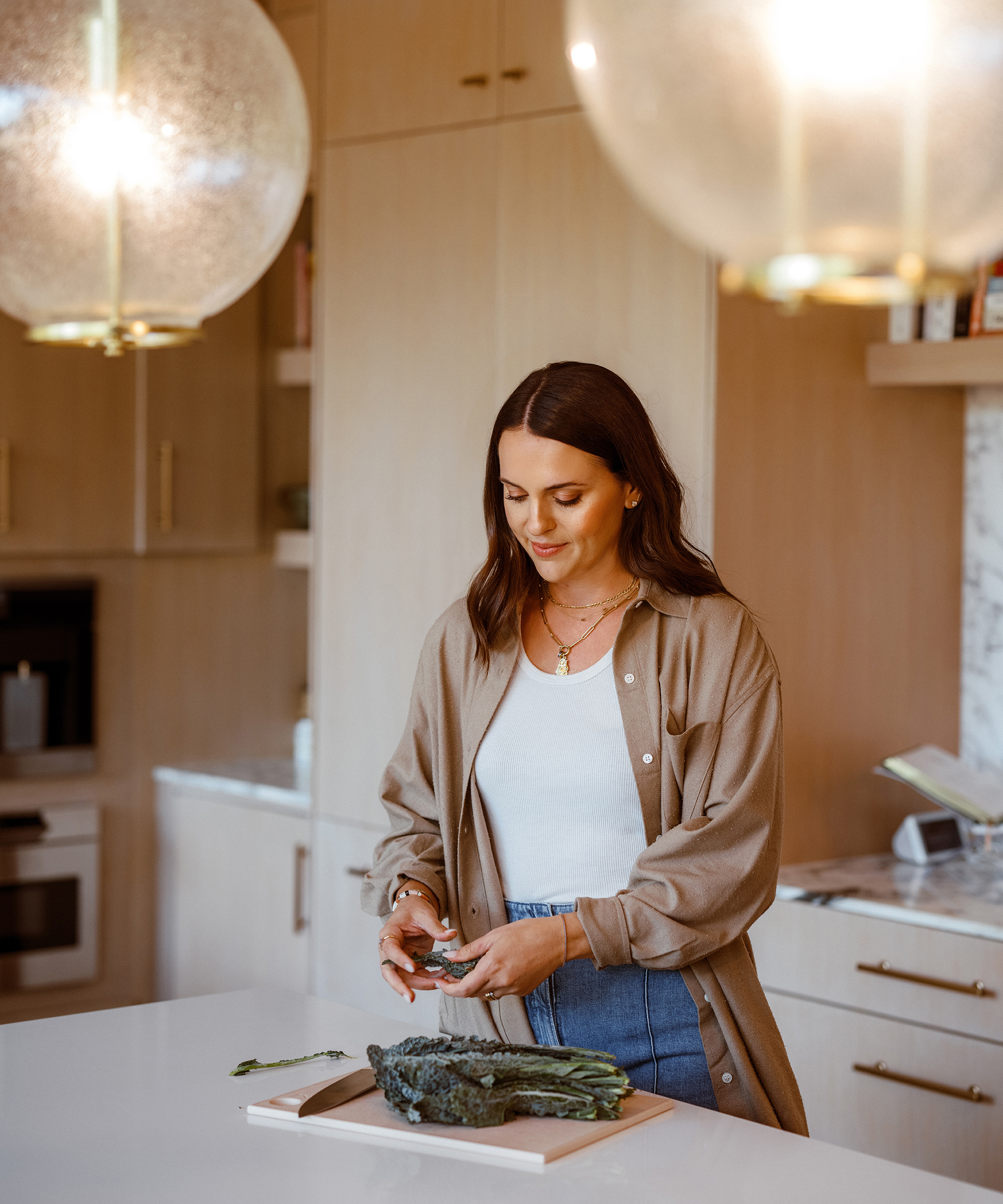 A woman, reminiscent of Alex Snodgrass, stands in a modern kitchen, preparing leafy greens on a cutting board. She wears a beige shirt over a white top and jeans, smiling as two round pendant lights hang above the countertop.