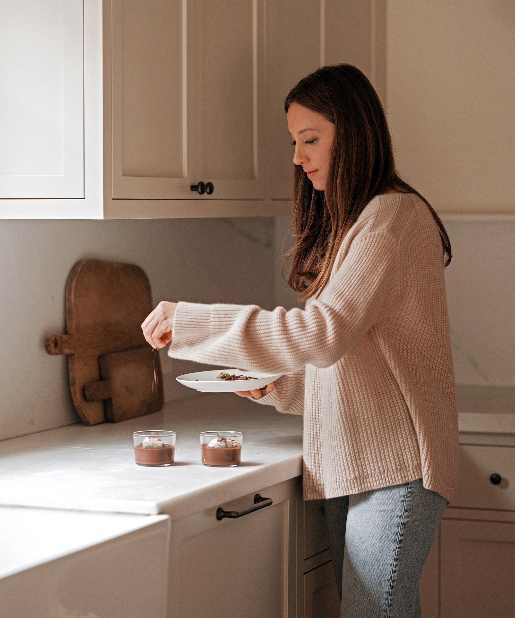 danielle in a cashmere boatneck sweater and jeans adding chocolate shavings to pot de creme dishes in a white kitchen