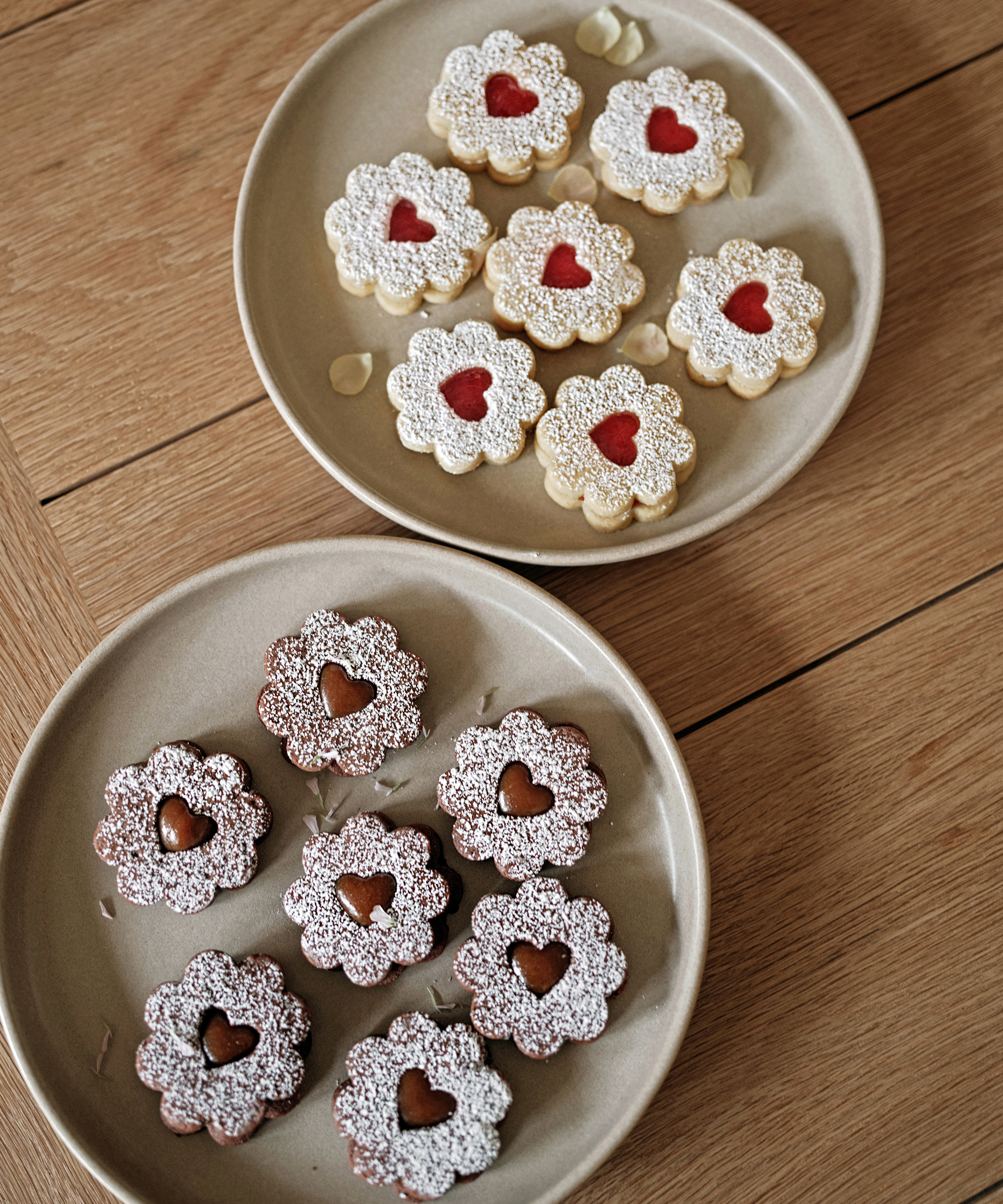 Two plates of flower-shaped sandwich cookies on a wooden table—perfect valentine desserts. The top plate holds light cookies with red jam centers, while the bottom has darker cookies with chocolate centers, all dusted with powdered sugar.