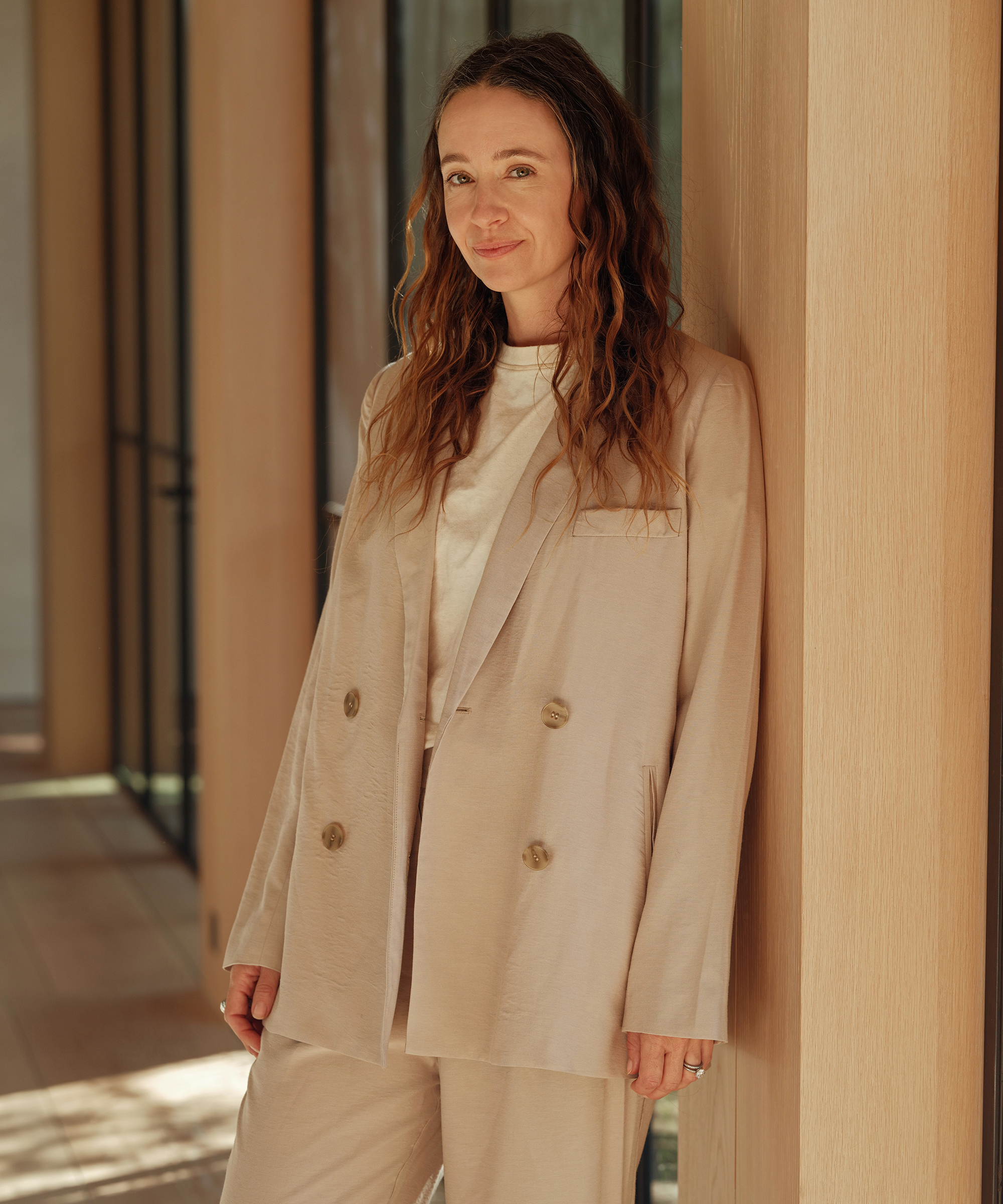jenni in a tan blazer with a white tee leaning against a wooden wall
