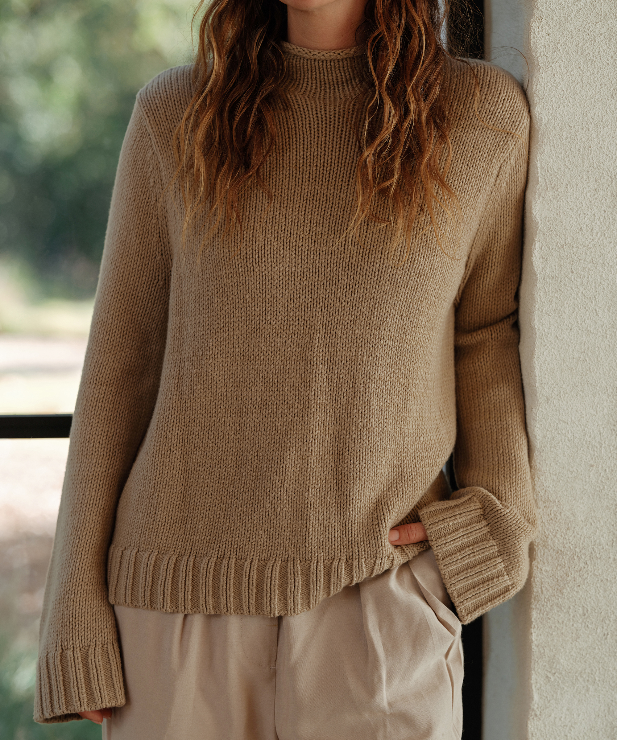 jenni in a taupe mockneck pullover with her hand in the pocket of her trousers leaning against an ivory brick wall