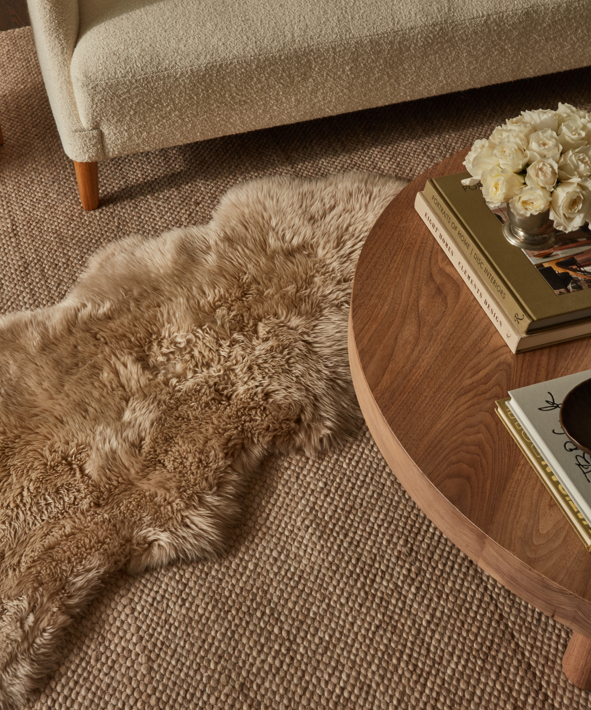 A cozy living room corner with a textured beige rug, a tan faux fur throw, a round wooden coffee table holding books and a bouquet of white roses—perfect for a relaxing Valentine’s Day—next to part of a cream upholstered sofa.