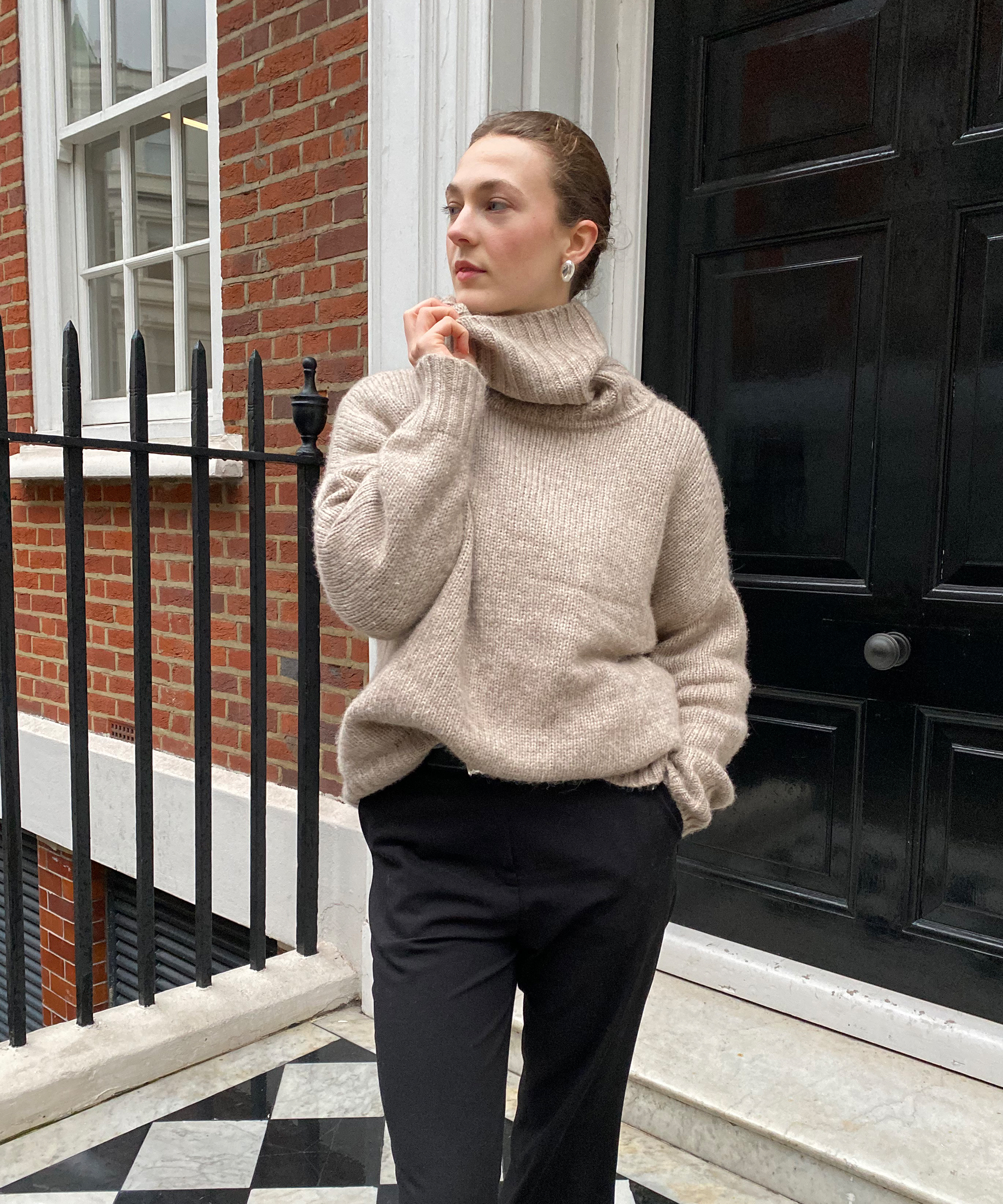 A person showcases street style on a checkered tile step in front of a black door and brick building, wearing a beige turtleneck sweater and black pants, with one hand in their pocket and the other touching their chin.