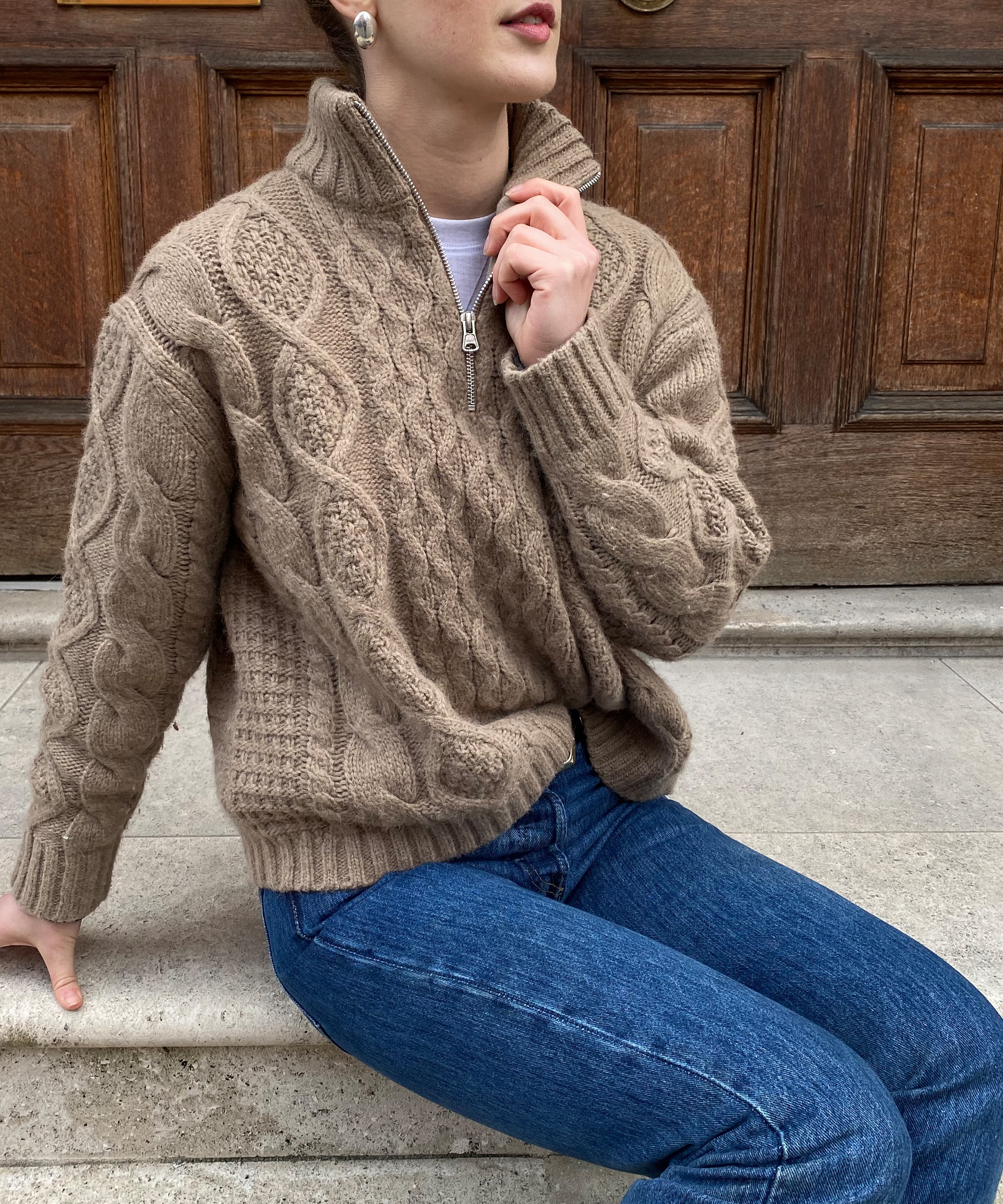 a woman sitting on stone steps in a taupe cable knit half zip and blue jeans
