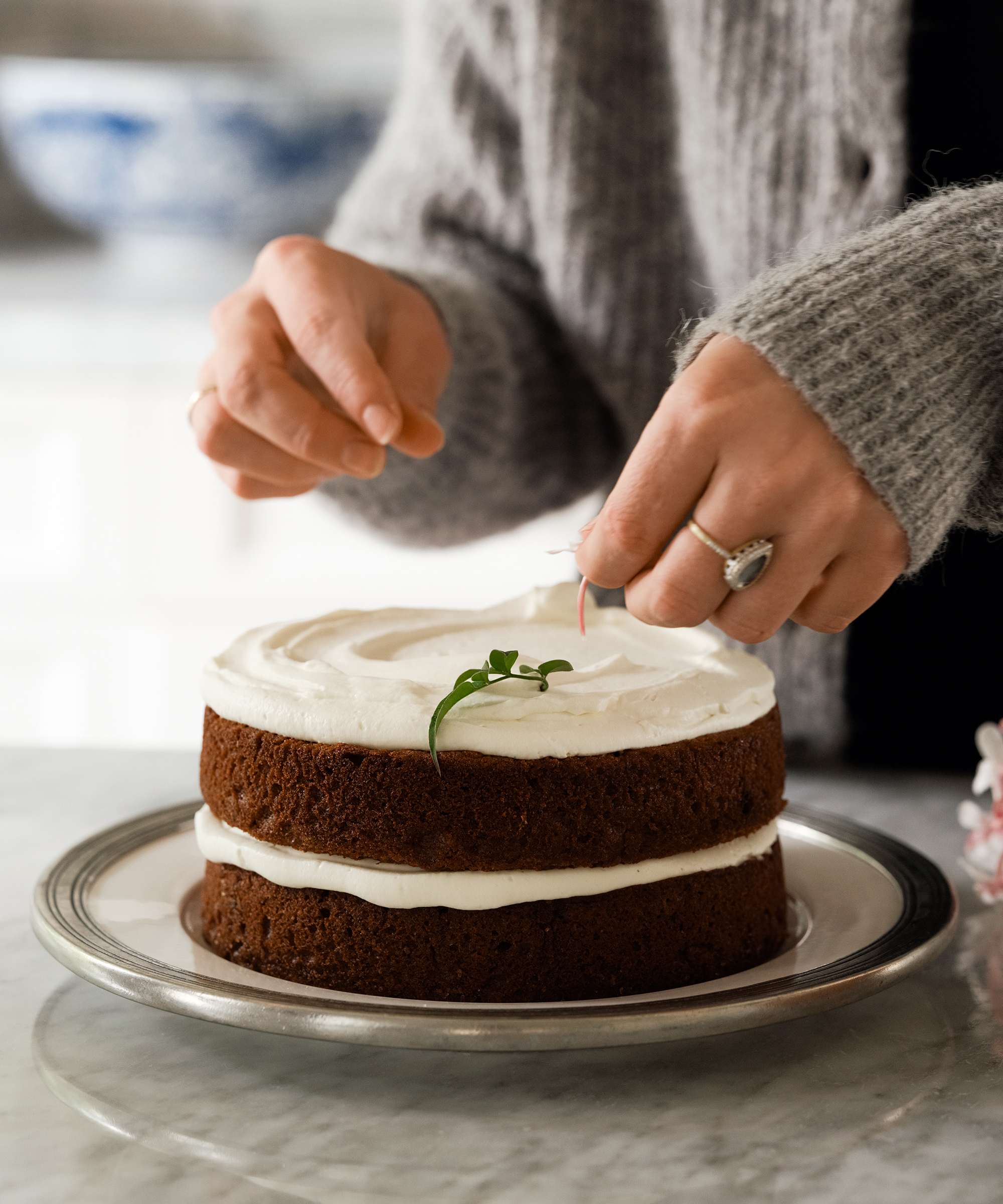 A person in a gray sweater decorates a two-layer olive oil cake with white frosting and greenery, placing a small garnish on top while the cake sits on a silver platter.