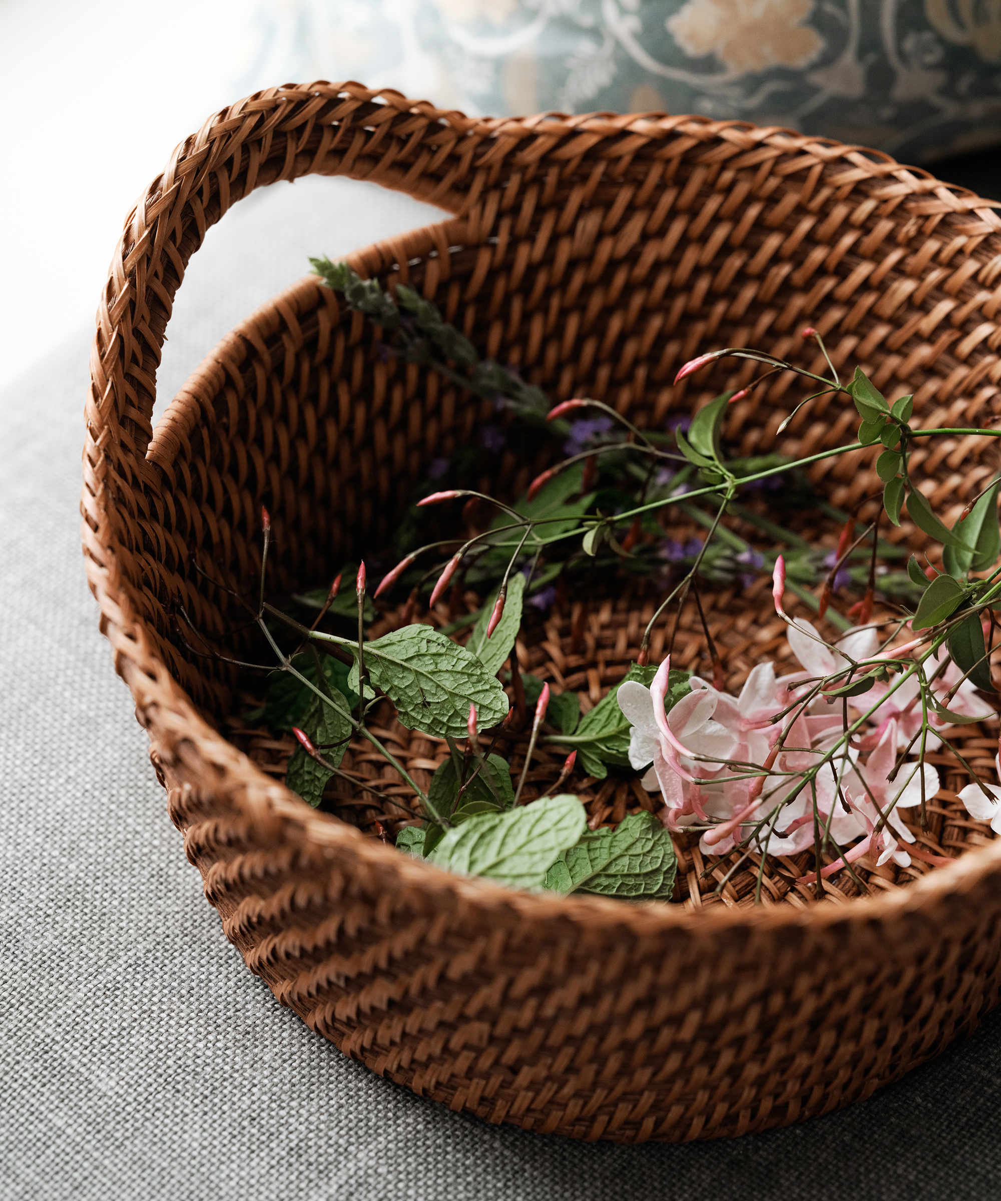 A woven basket containing sprigs of lavender, green leaves, and small pink and white flowers, reminiscent of a charming olive oil grove, resting on a gray fabric surface.