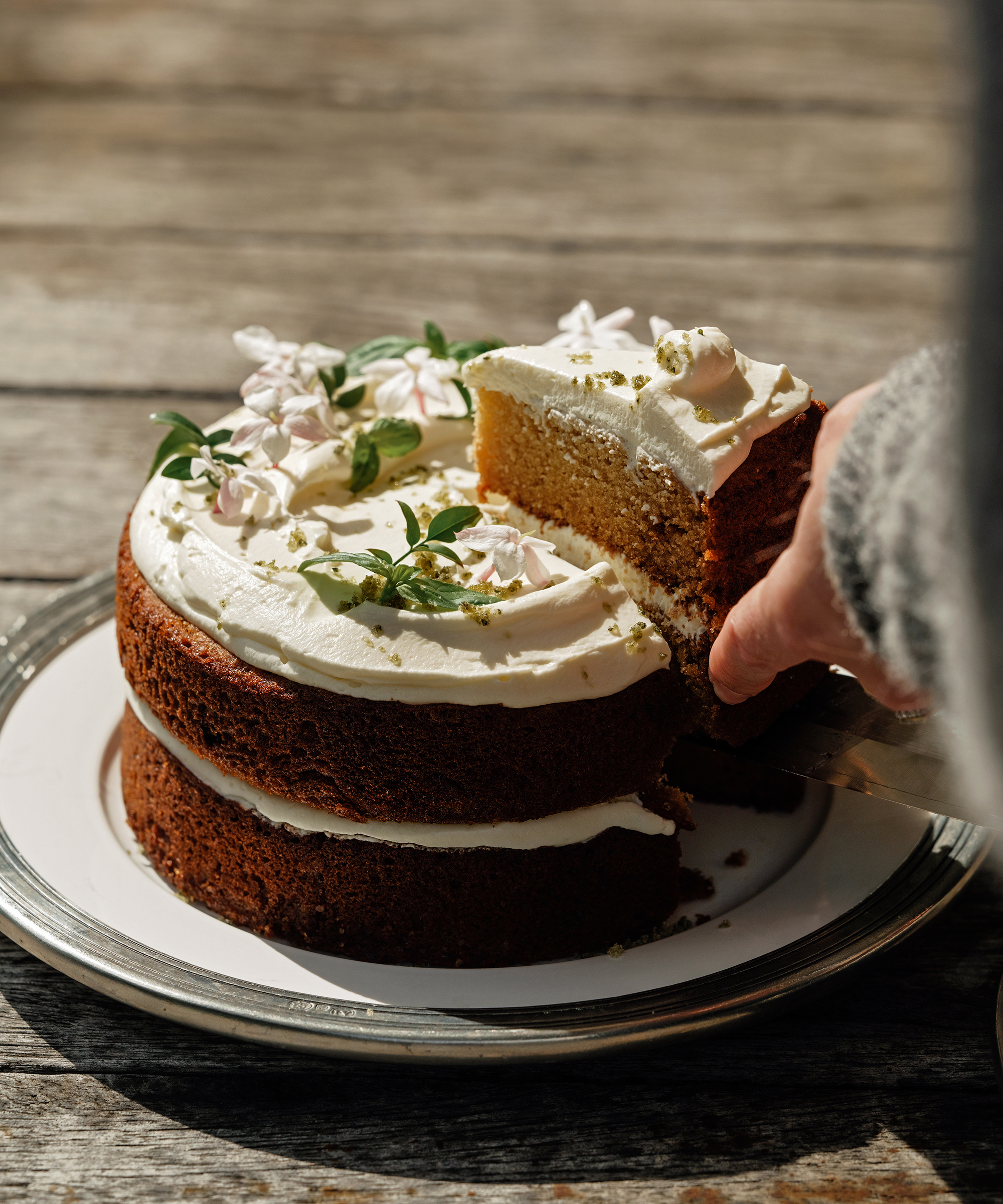 A hand lifts a slice from a two-layer olive oil cake with white frosting, green leaves, and small white flowers on top, displayed on a white plate and silver tray on a rustic wooden table.