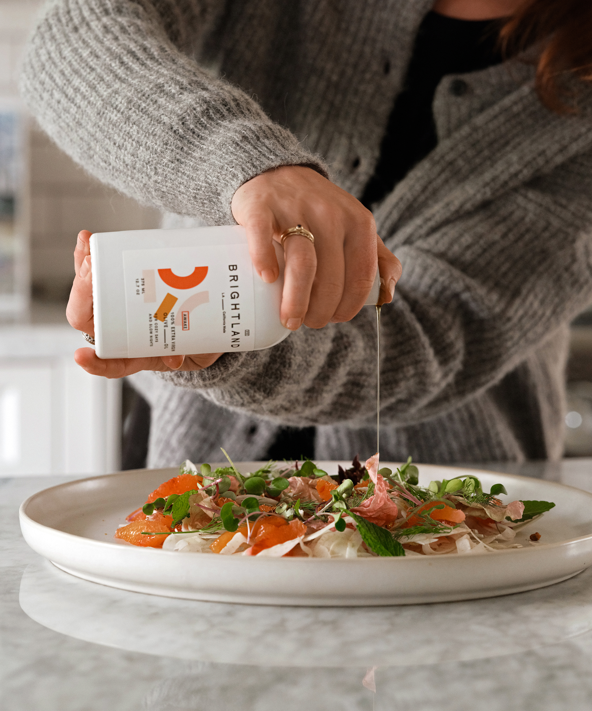 A person in a gray sweater pours olive oil dressing from a Brightland bottle onto a salad with greens, citrus slices, and nuts on a white plate atop a marble countertop.