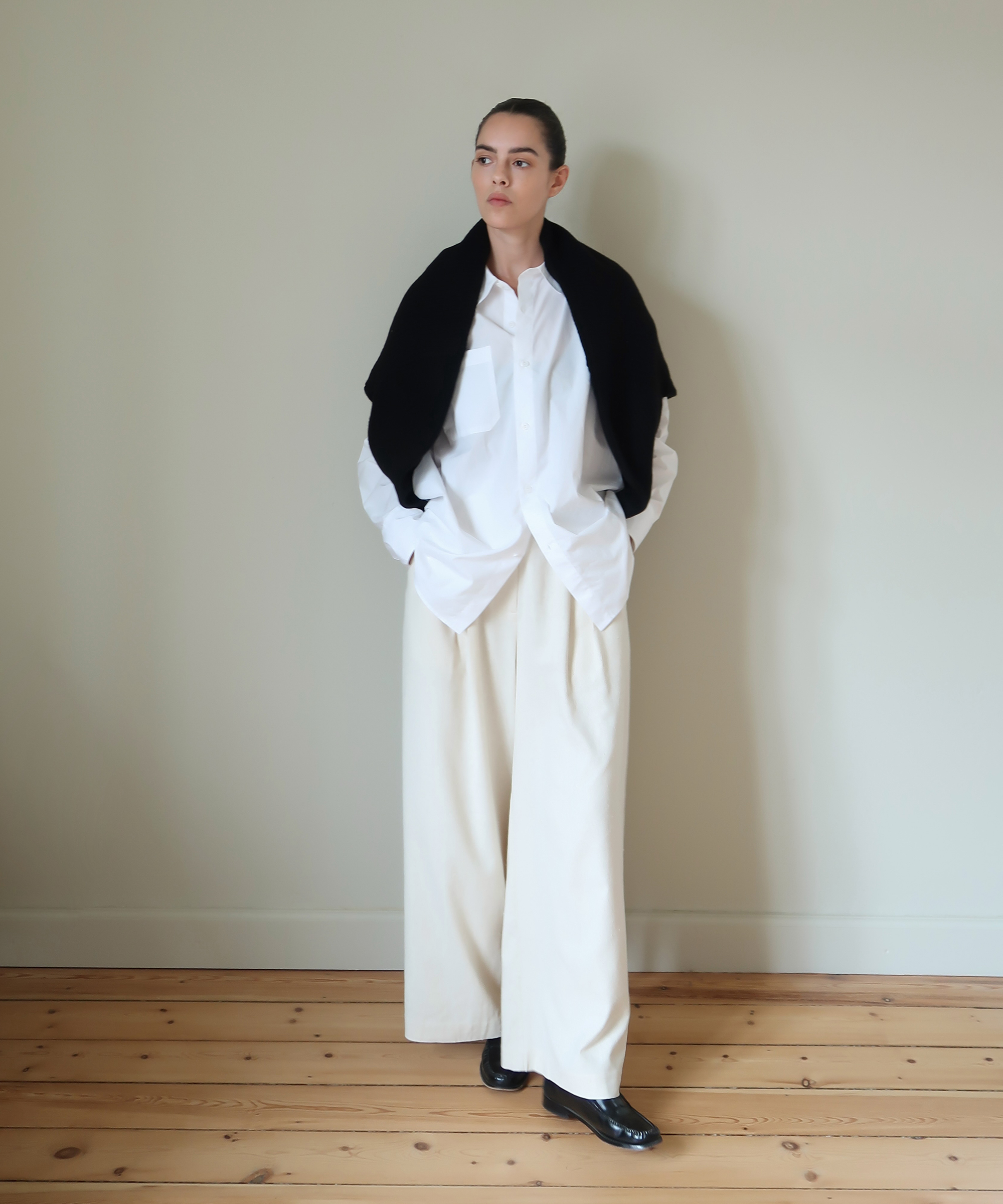 A person stands indoors against a plain wall, showcasing effortless spring style in a white oversized shirt, wide-leg cream trousers, black shoes, and a black sweater draped over their shoulders. Hands in pockets, they look thoughtfully to the side.