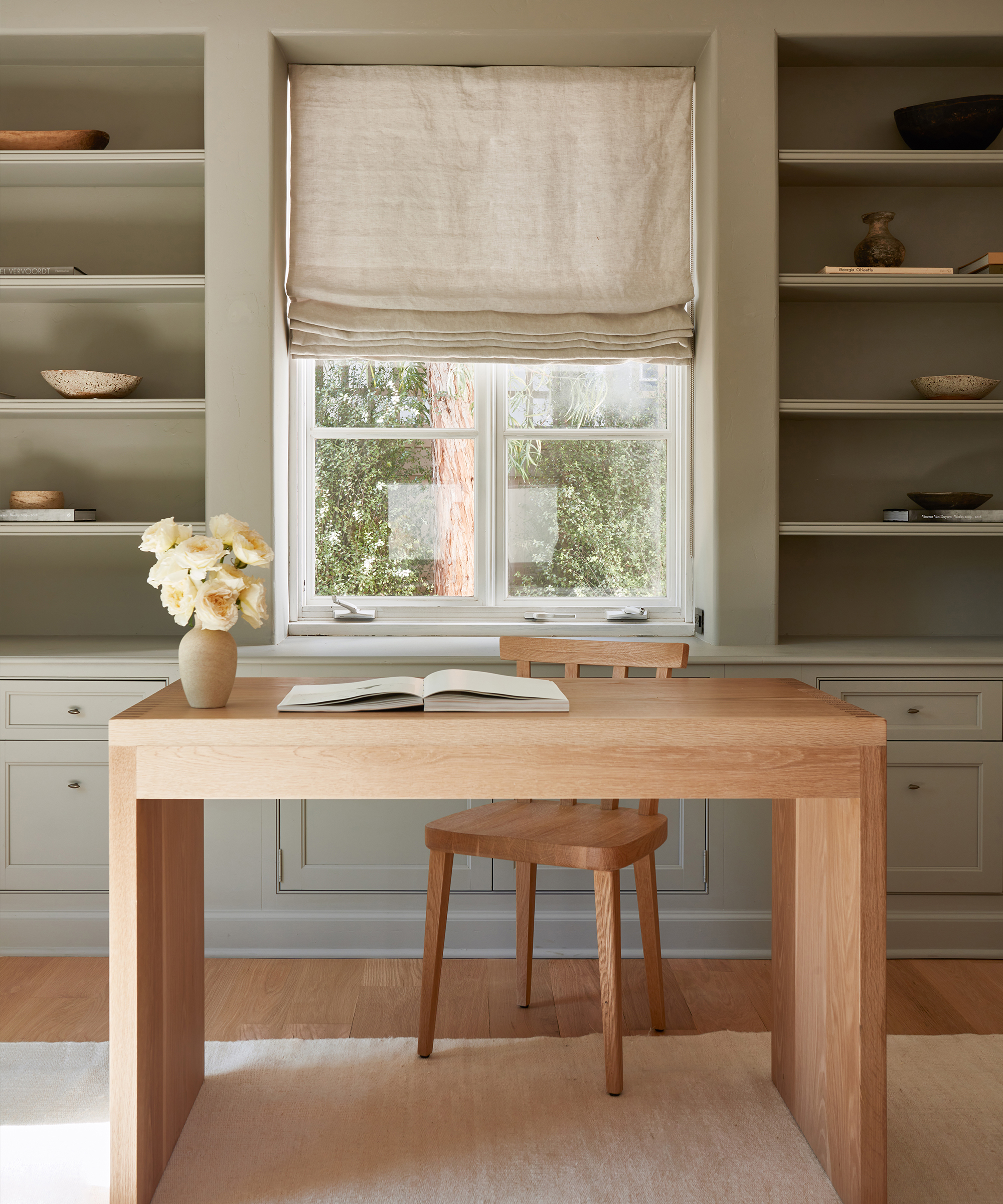 A minimalist home office with a wooden desk and chair, open book, vase of white flowers, built-in shelves with decor, and a window featuring linen Roman shade window treatments that welcome natural light.