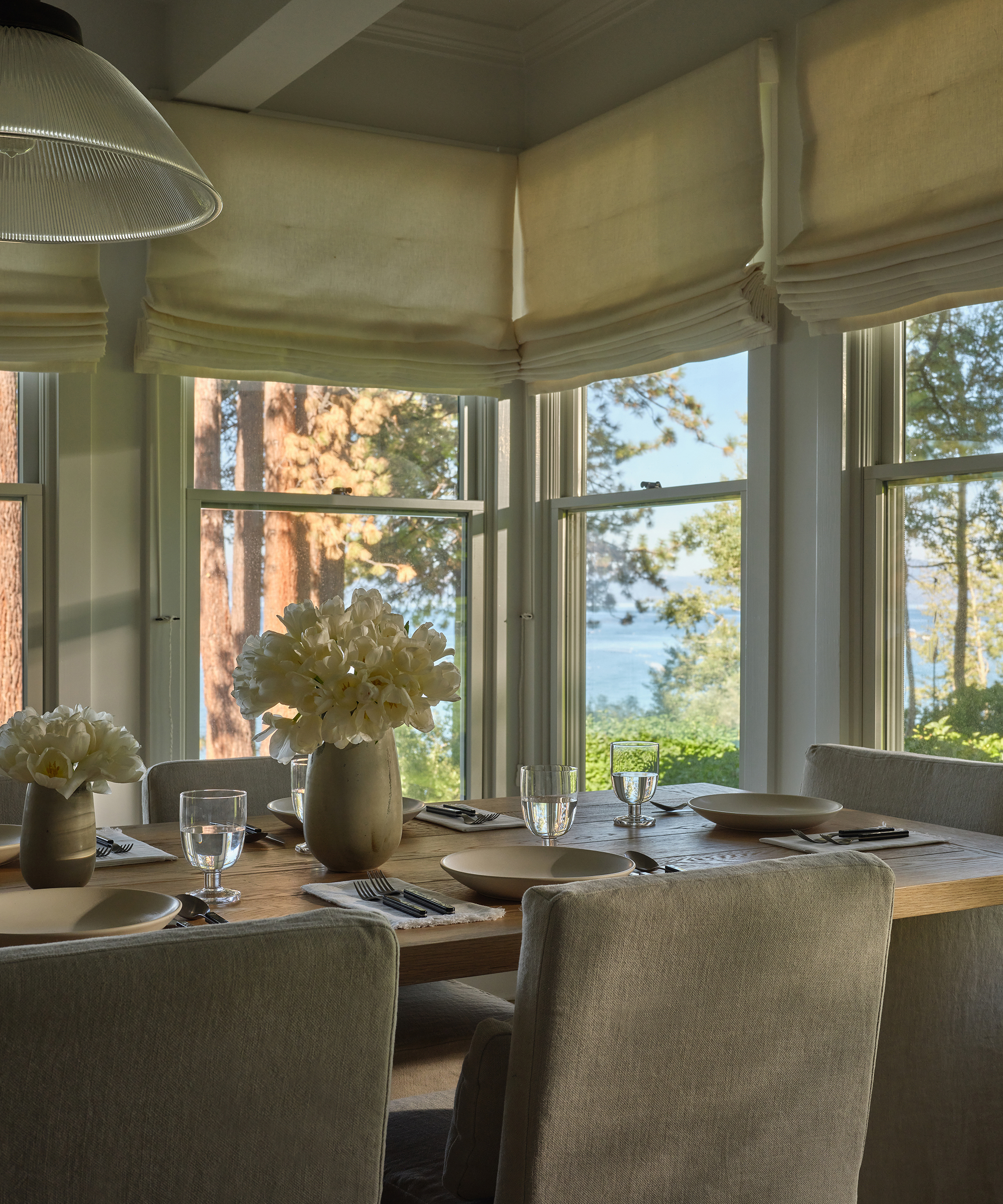 A sunlit dining area with beige upholstered chairs, a wooden table set with plates, glasses, and neutral vases of white flowers sits by large windows adorned with elegant window treatments, overlooking trees and a distant lake view.