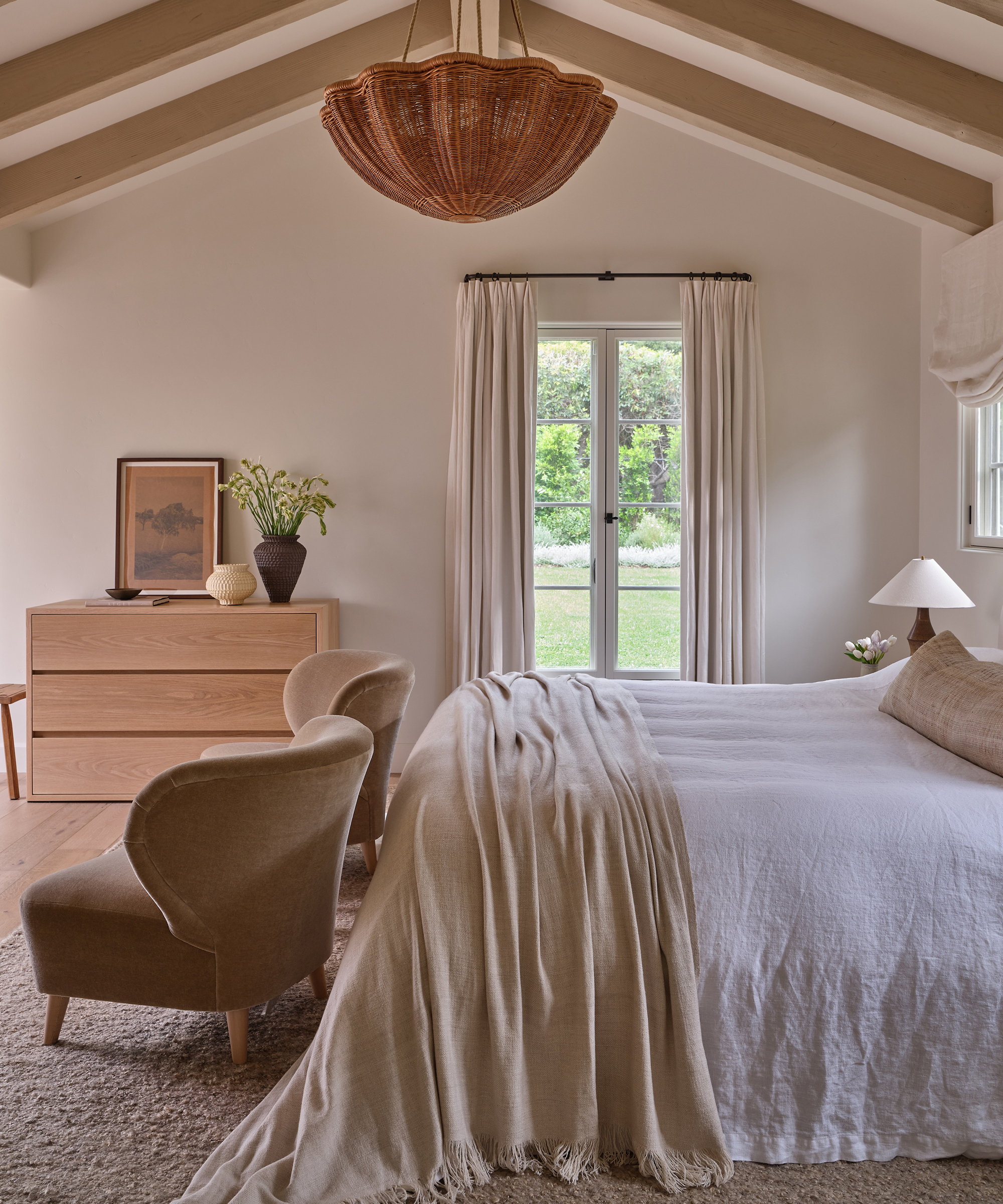 A cozy bedroom with a white bed and beige blanket, two beige armchairs, a wooden dresser with decor, elegant window treatments, and a large woven chandelier. Sunlight streams in through glass doors overlooking greenery.