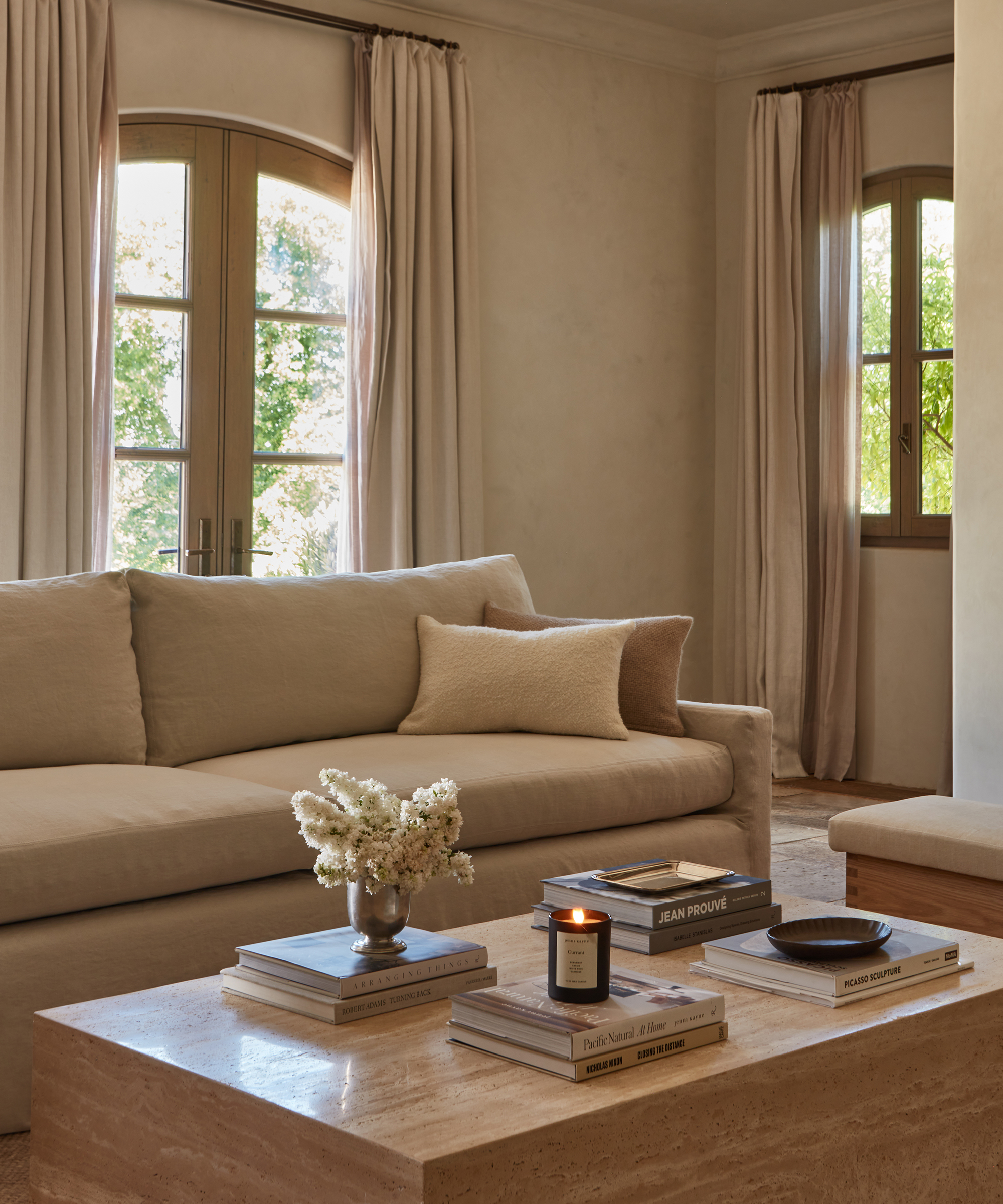 A cozy living room with a beige sofa, a light pillow, and elegant window treatments. A marble coffee table holds stacked books, a candle, and a small vase of white flowers as sunlight streams through arched windows.