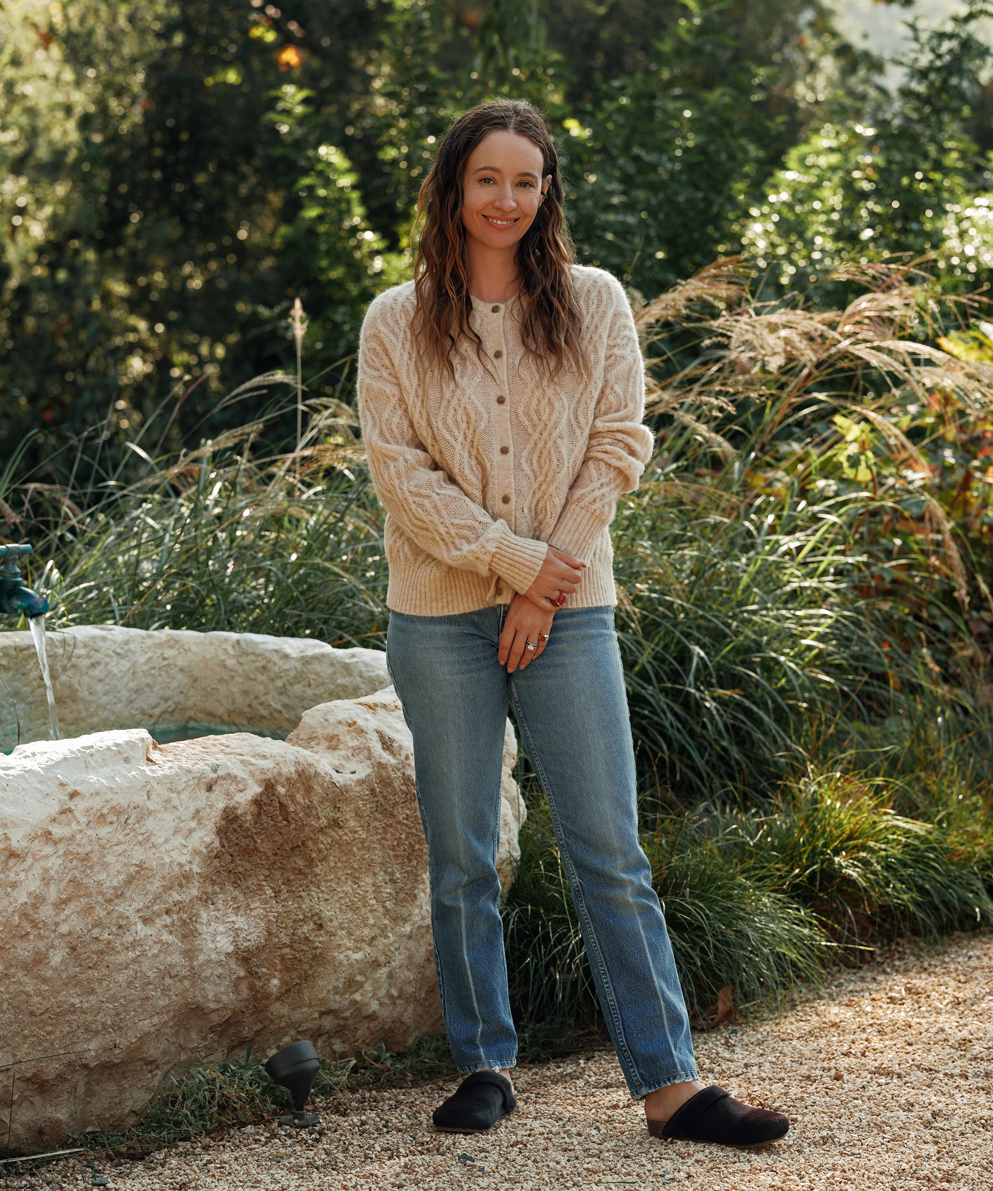 jenni in a sand cable nell cardigan with jeans and cowhide moccasins standing on gravel next to a water feature