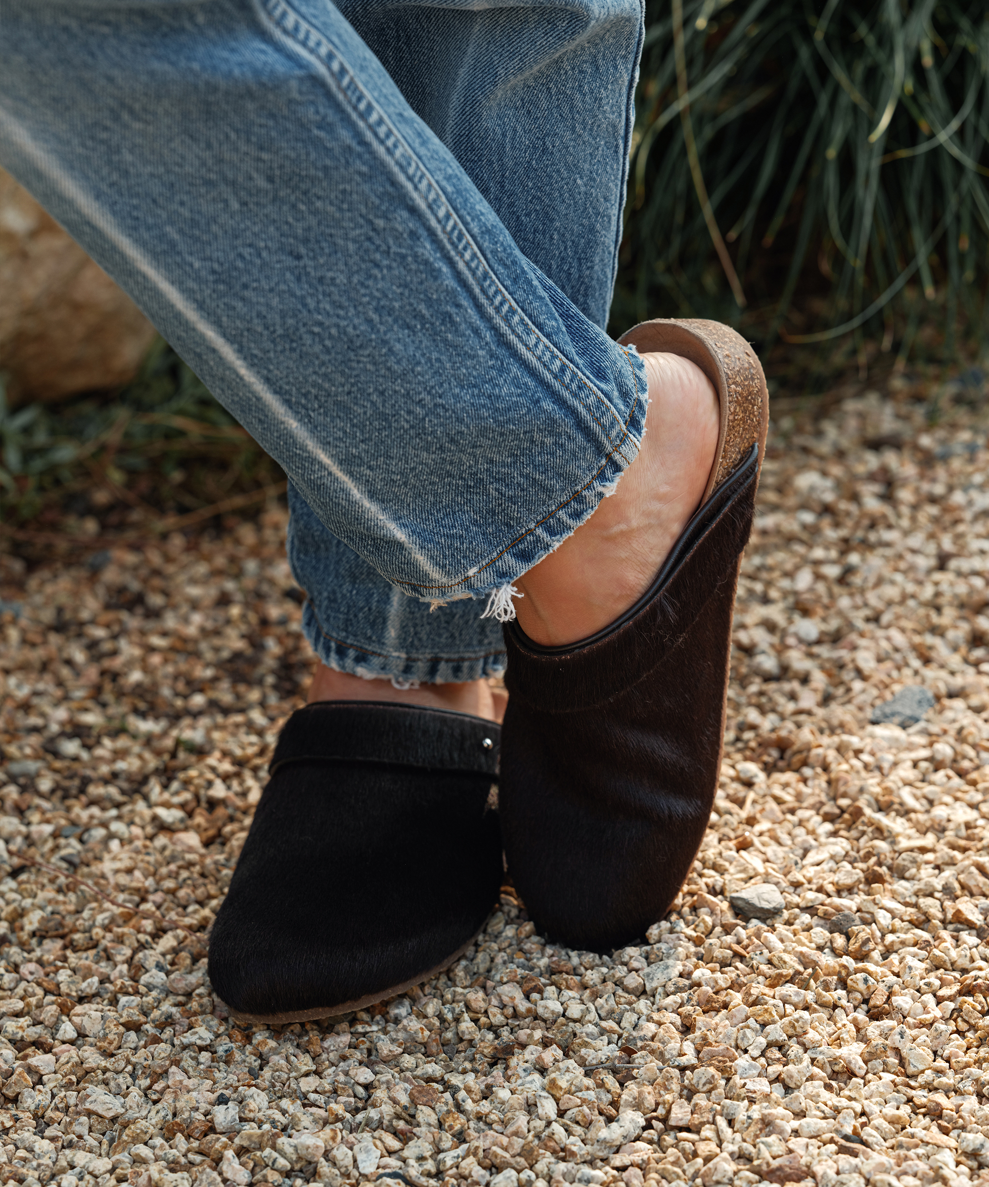 A person wearing blue jeans and black slip-on clogs with cork soles stands on a gravel surface, legs slightly crossed—a stylish choice for any midwinter wardrobe. Green plants are visible in the background.