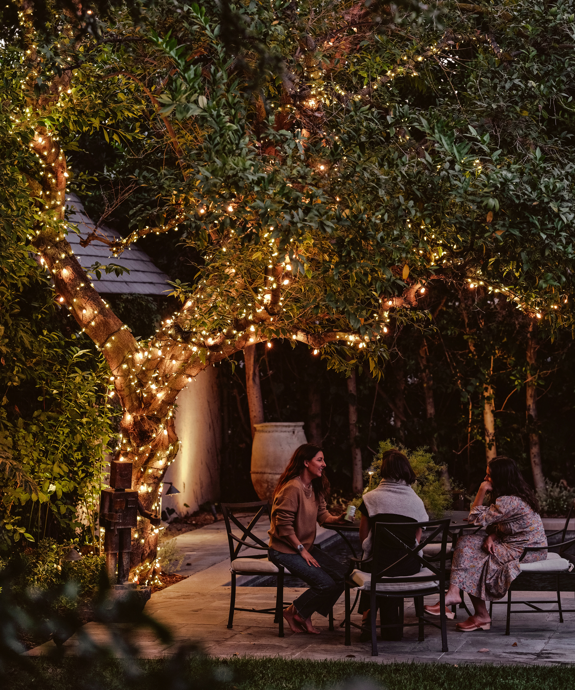 people sitting in outdoor accent chairs under a tree draper in lights in a backyard