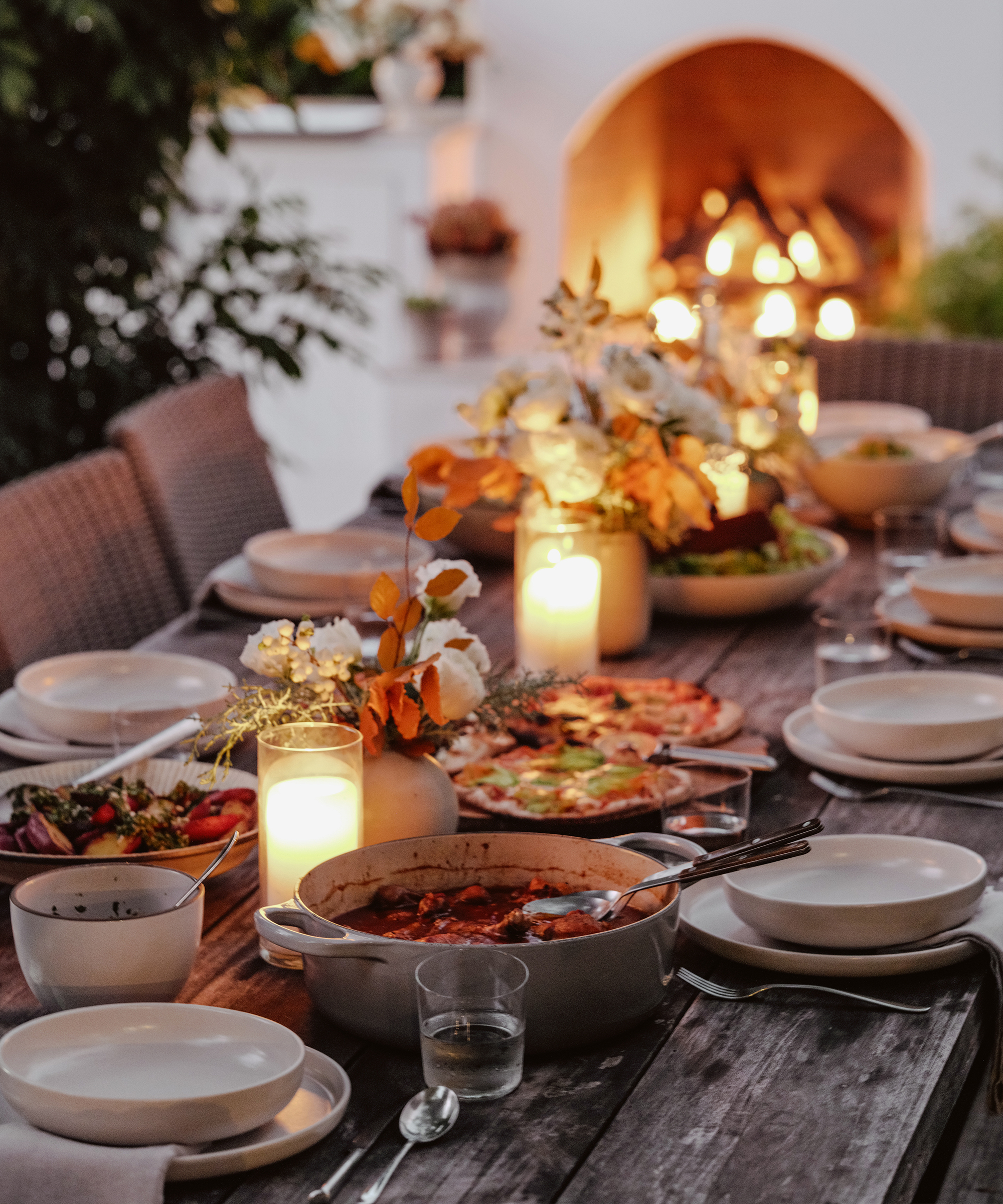 an outdoor dining table set with taupe plates linen napkins and candles leading to an outdoor fireplace
