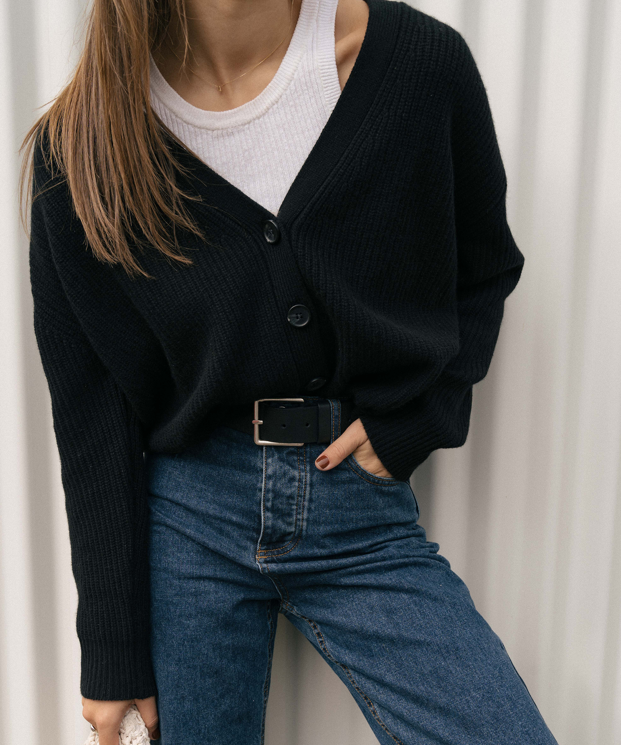 a woman standing in front of an ivory wall in a jeans and a navy sweater with a white tank and a black belt