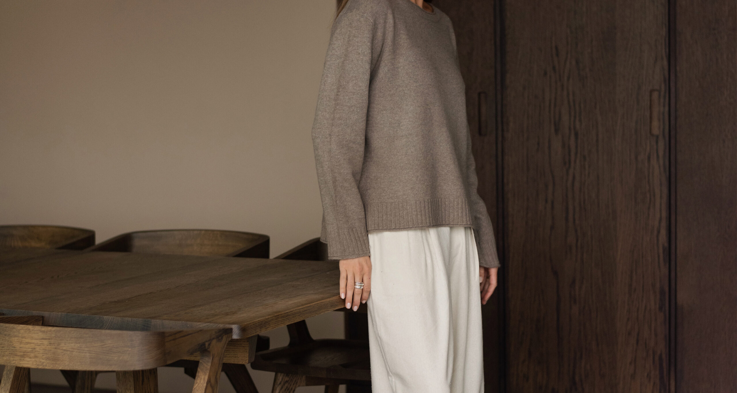 A person showcases a spring style in a taupe sweater and white pants, standing beside a wooden table and chairs, with a dark wooden cabinet in the background. Their face is not visible.