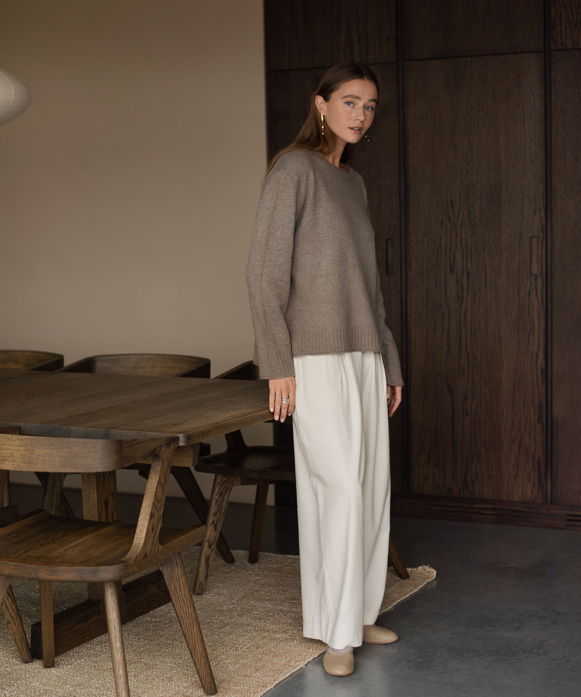 a woman in a taupe sweater and ivory trousers standing next to a wooden dining table and dining chairs