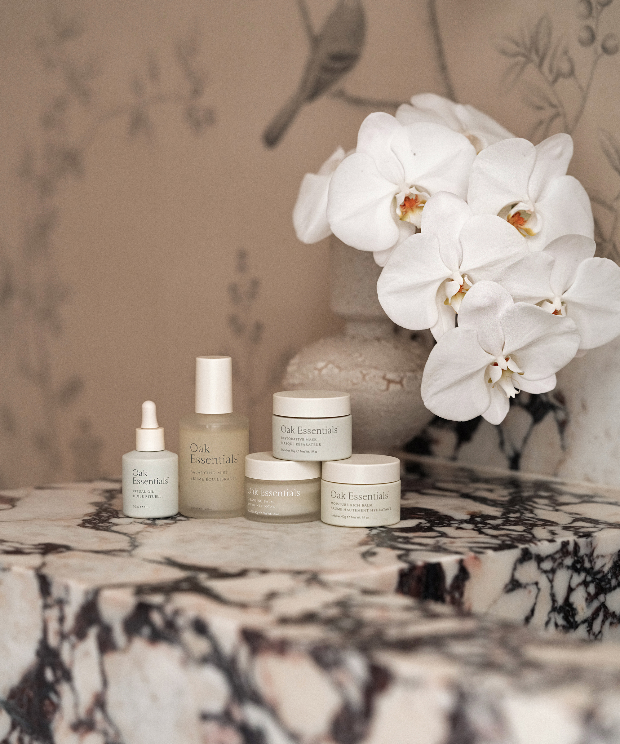 A collection of Oak Essentials skincare products is arranged on a marble surface next to a white orchid arrangement and decorative vase, evoking the serene elegance of Pacific Palisades against a beige, botanical-themed background.