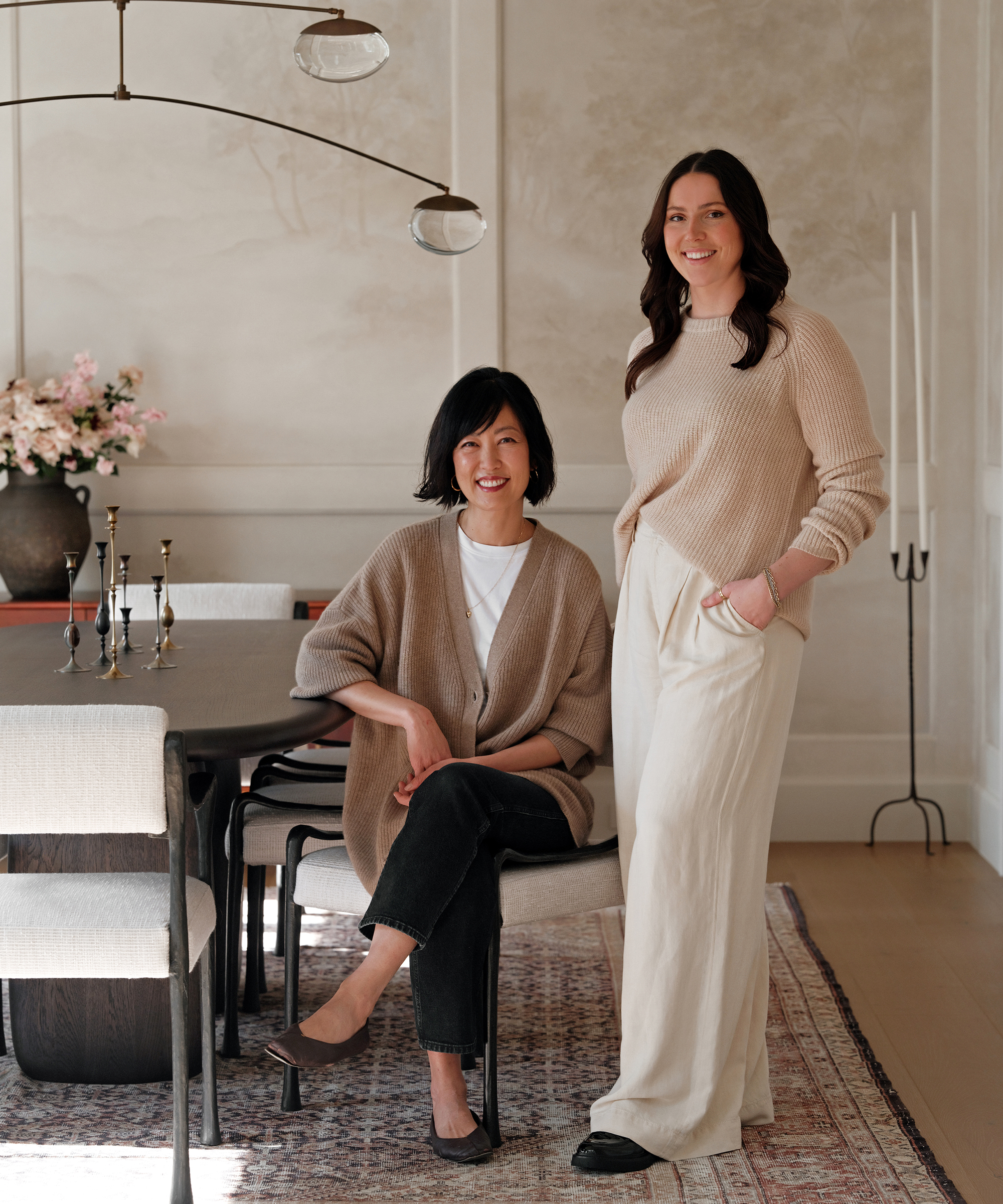 Two women pose in a stylish, softly lit Pacific Palisades dining room. One sits at the table in a beige cardigan and black pants, while the other stands beside her in a beige sweater and cream pants. A vase of flowers and candles are visible.