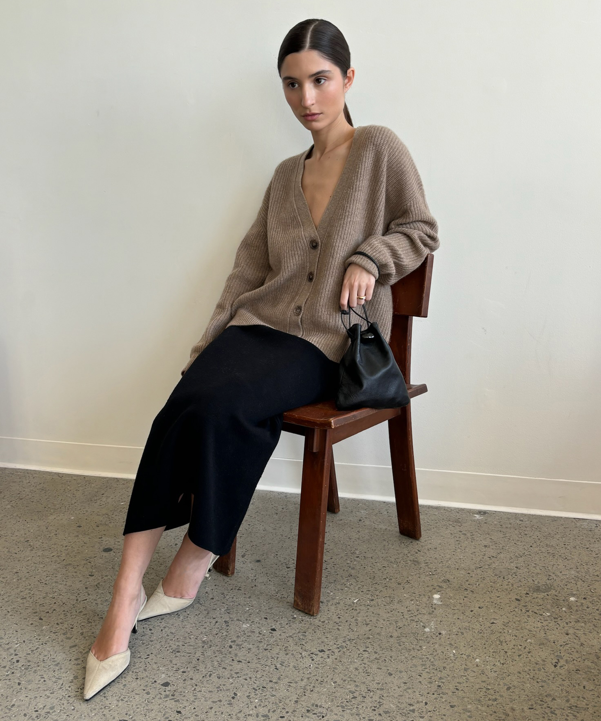 A woman with dark hair in a low bun sits on a wooden chair against a plain white wall. She wears a loose tan cardigan, black skirt, cream pointed heels, and holds a small black bag.