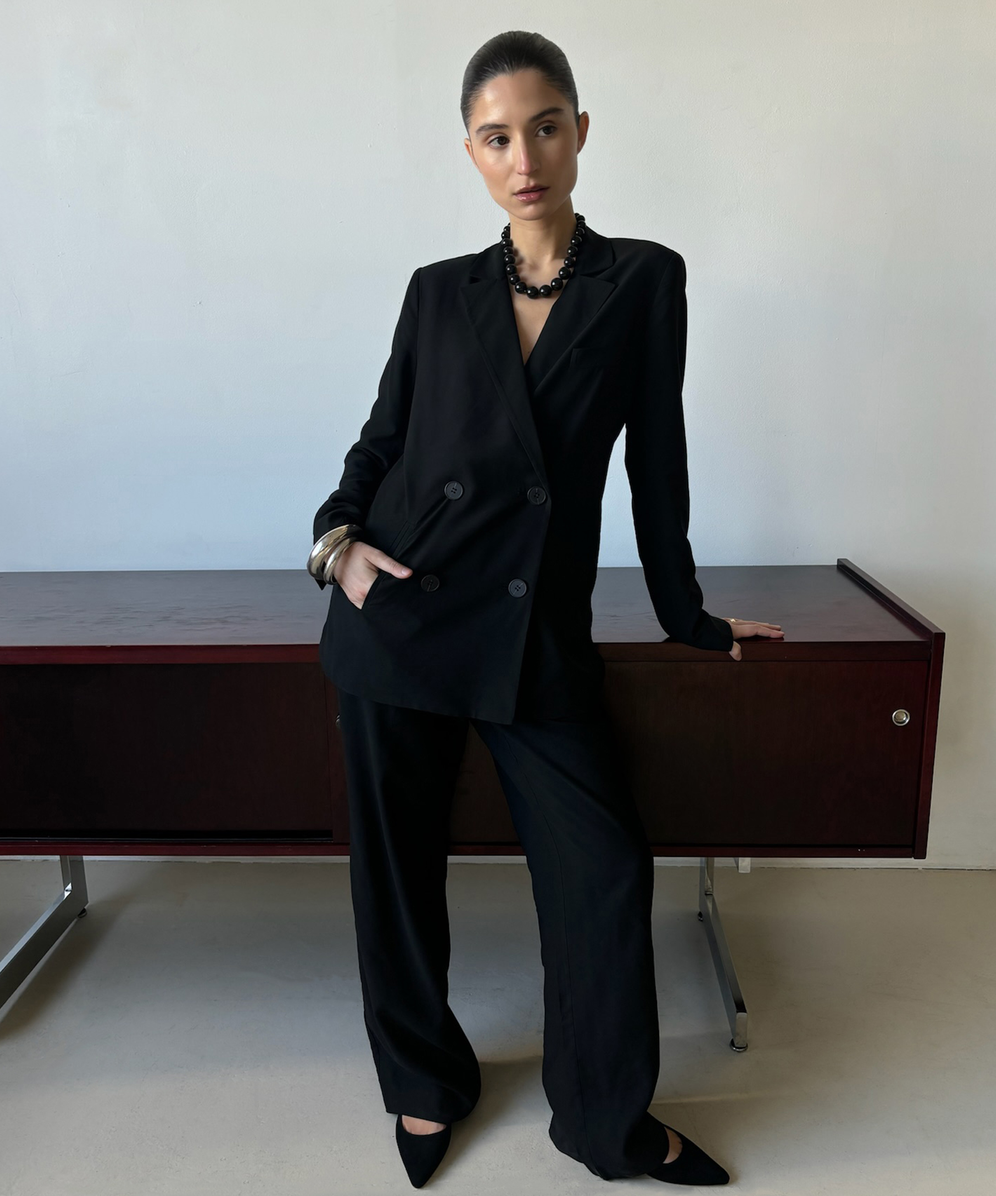 andrea standing next to a wood sideboard in a black blazer and matching black trousers with a kitten heel