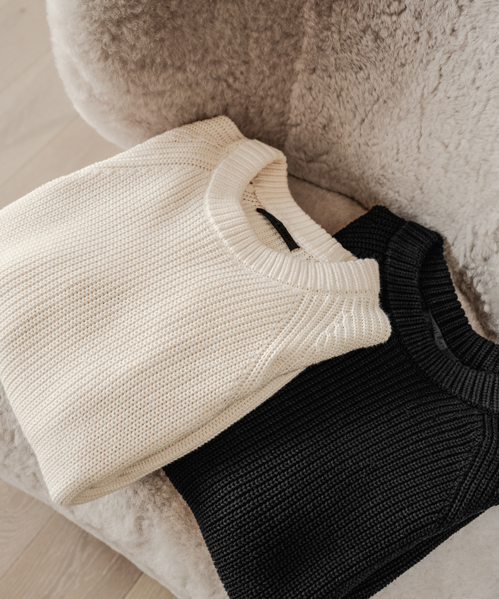 Two cozy knit sweaters, one cream-colored and one black, are placed side by side on a soft, beige, fuzzy chair.