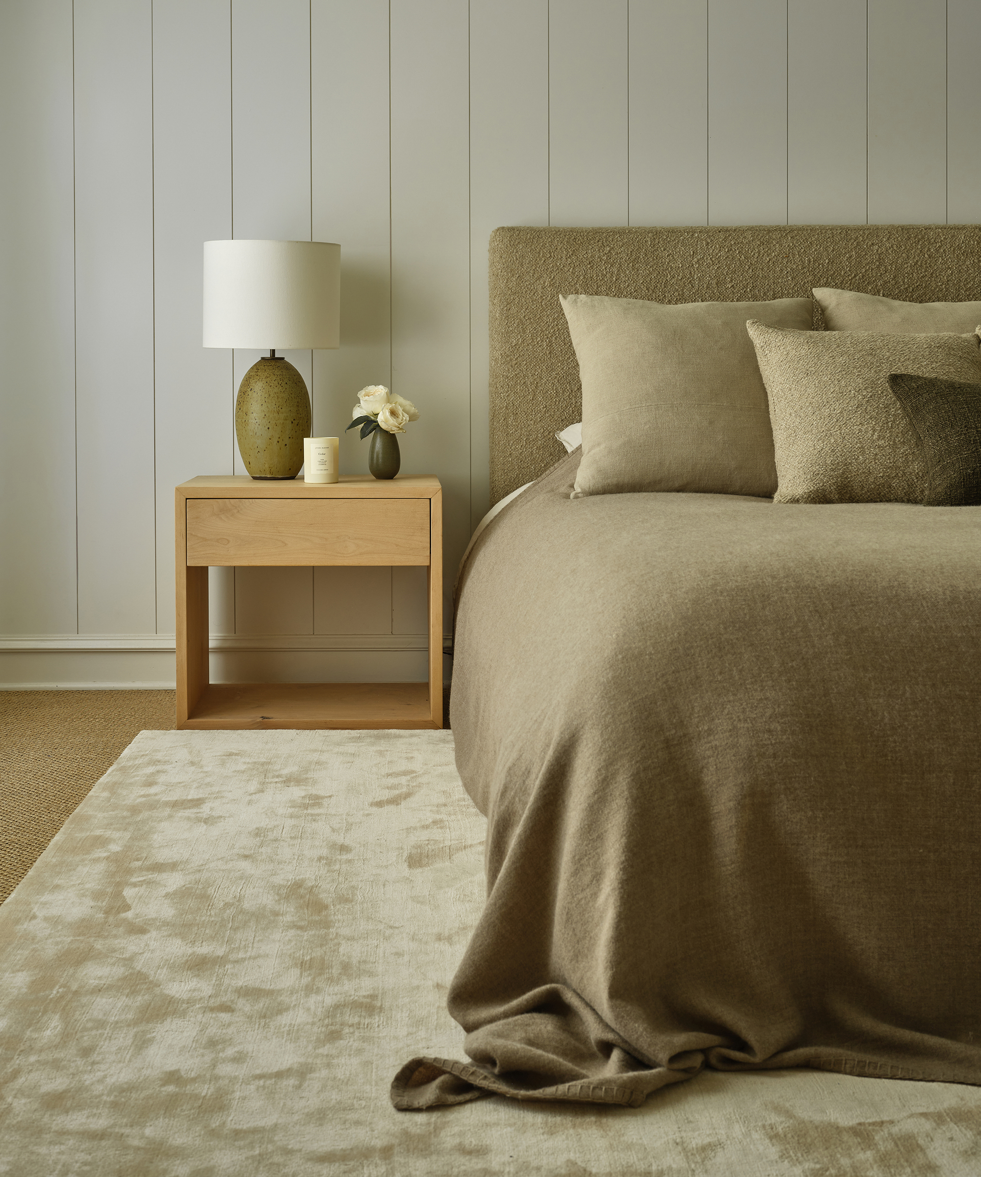 a bedroom with a plush area rug and a boucle bed with a taupe coverlet and a wooden nightstand with a ceramic lamp