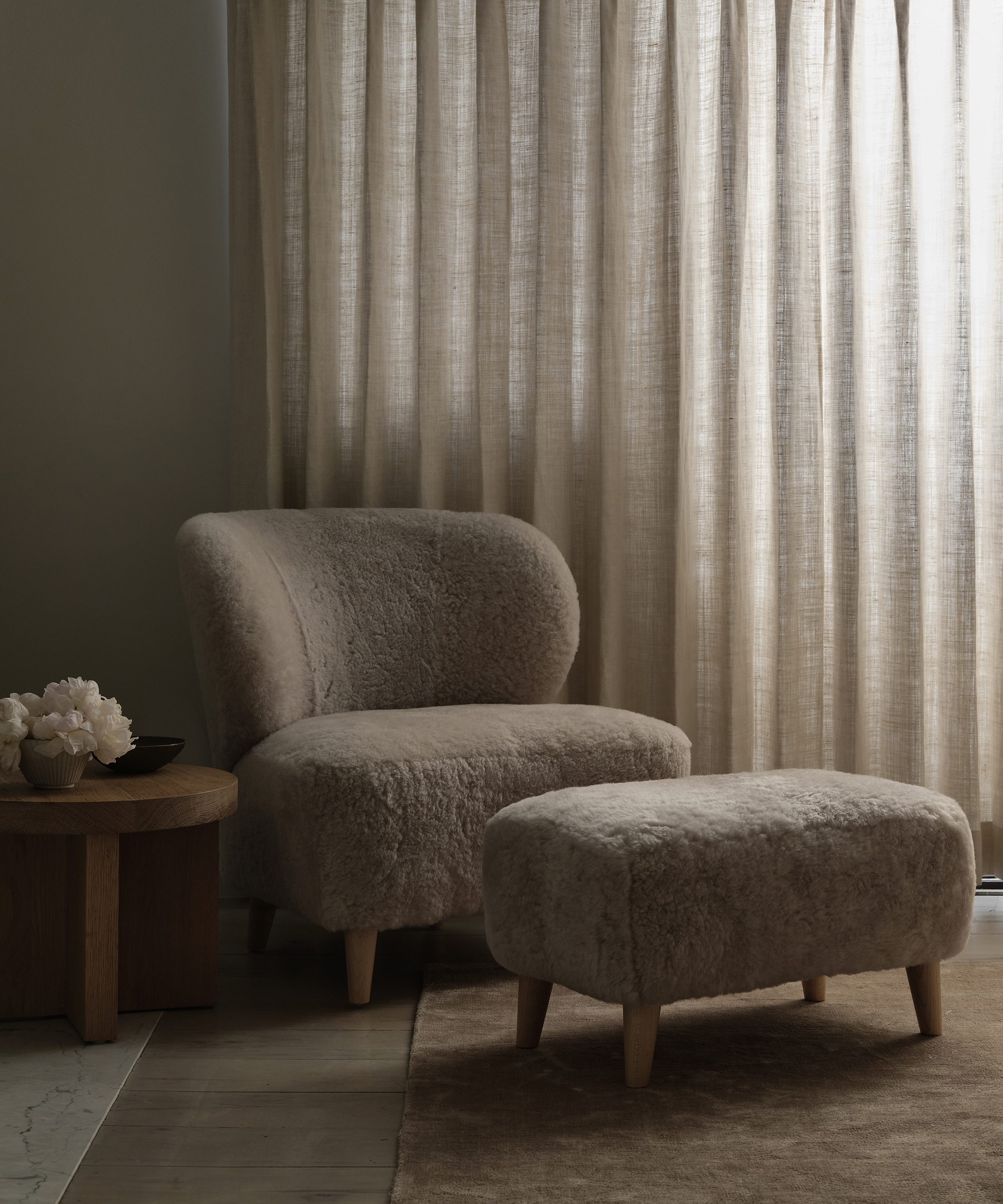 A cozy, budgeting-friendly chair and matching ottoman with soft, textured fabric sit on a rug next to a round wooden side table, in front of floor-length beige curtains in a softly lit room.