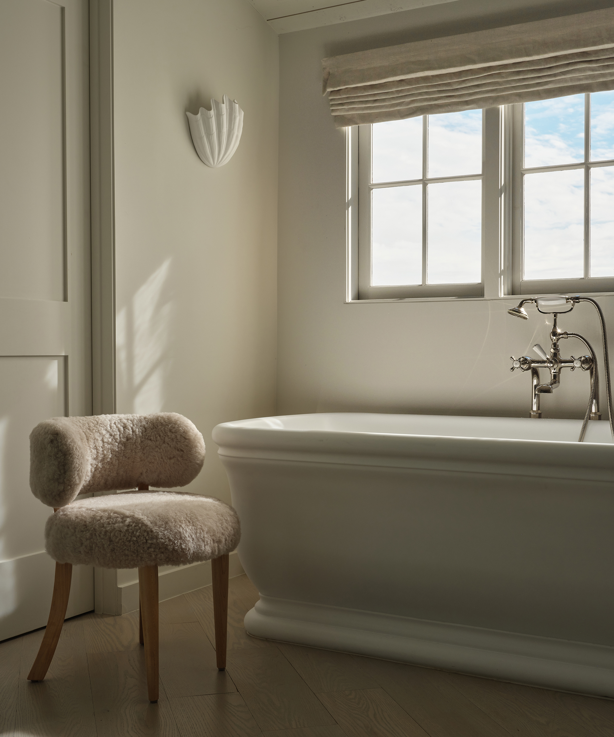 Sunlight streams into a minimalist, budgeting-friendly bathroom with a white freestanding tub, a plush furry chair with wooden legs, a shell-shaped wall sconce, and a window with beige roman shades revealing a partly cloudy sky.