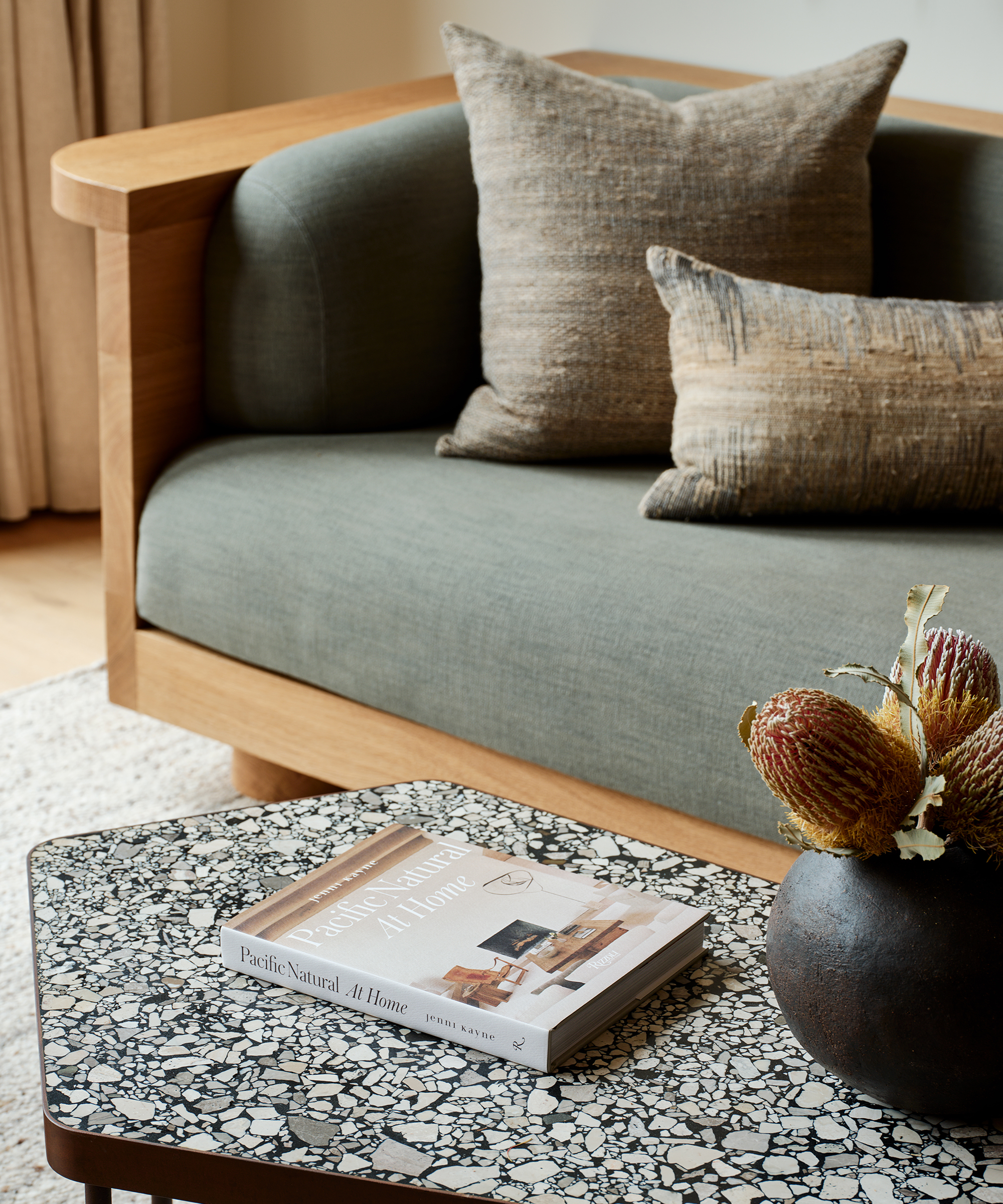 a terrazzo coffee table with a coffee table book next to a wooden and green upholstered couch