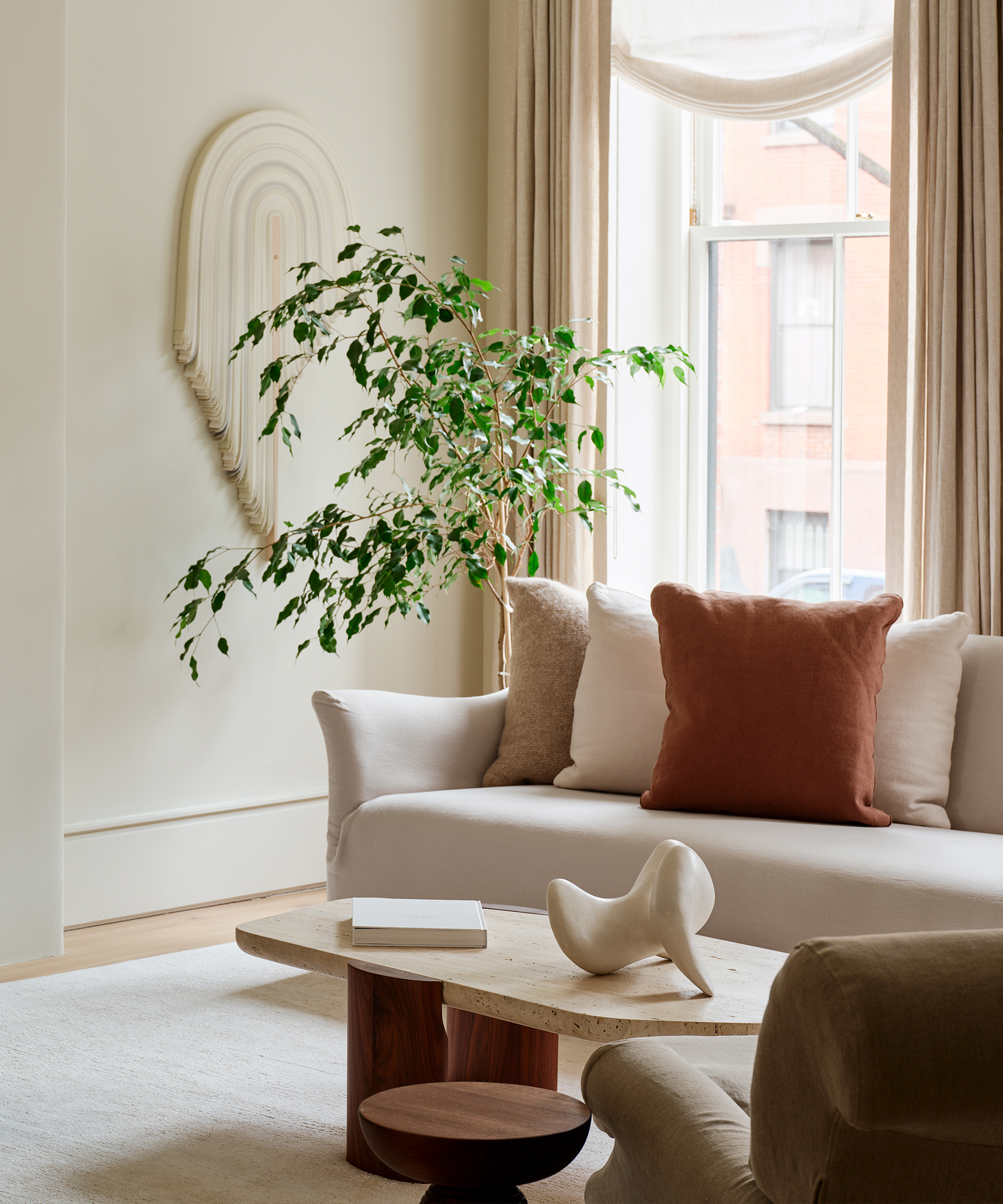 A modern living room in a Brooklyn brownstone with a light beige sofa, rust-colored and beige pillows, a sculptural coffee table, abstract wall art, a leafy plant, and large windows with sheer curtains letting in natural light.