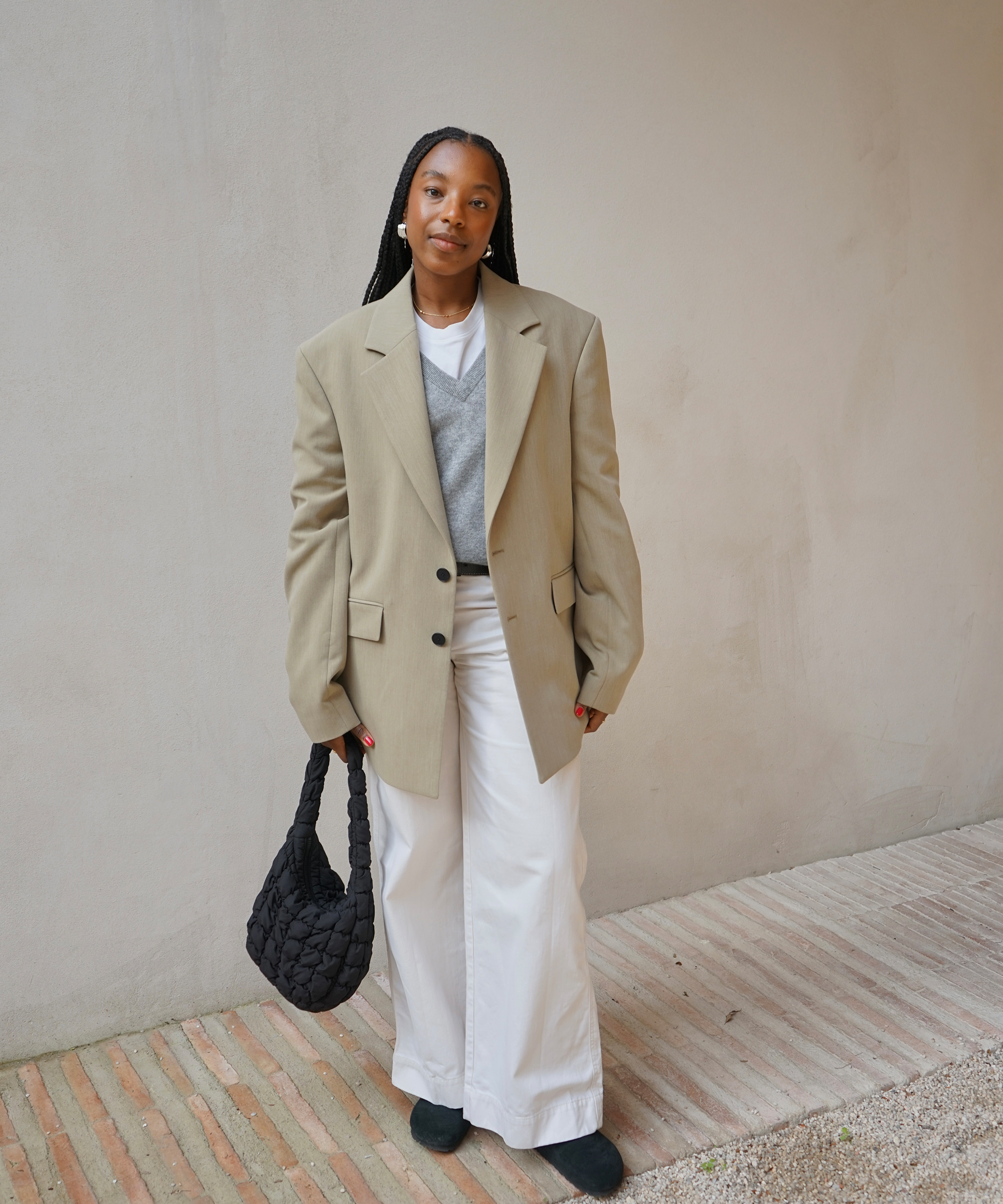 taffy standing in front of a neutral plaster wall wearing white jeans a tan blazer and grey vneck sweater