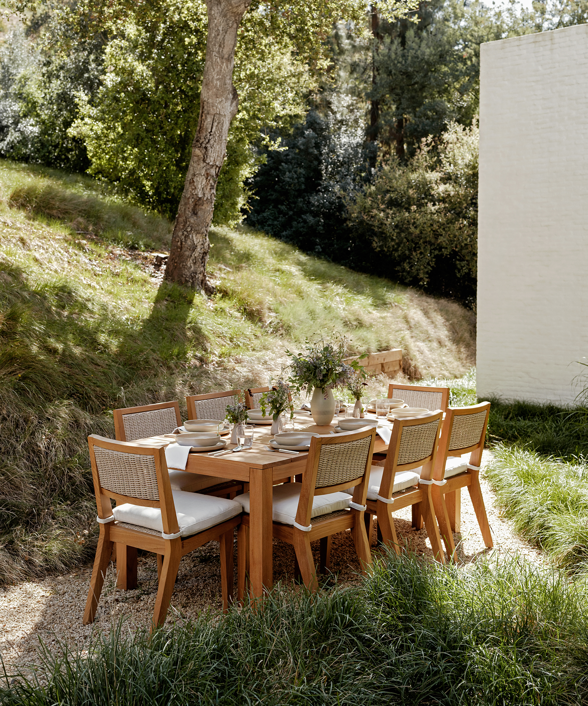 A wooden outdoor dining table with eight cushioned chairs is set for a spring dinner on a gravel patio, surrounded by lush green grass and trees beside a white brick building. A vase with flowers sits at the center of the table.