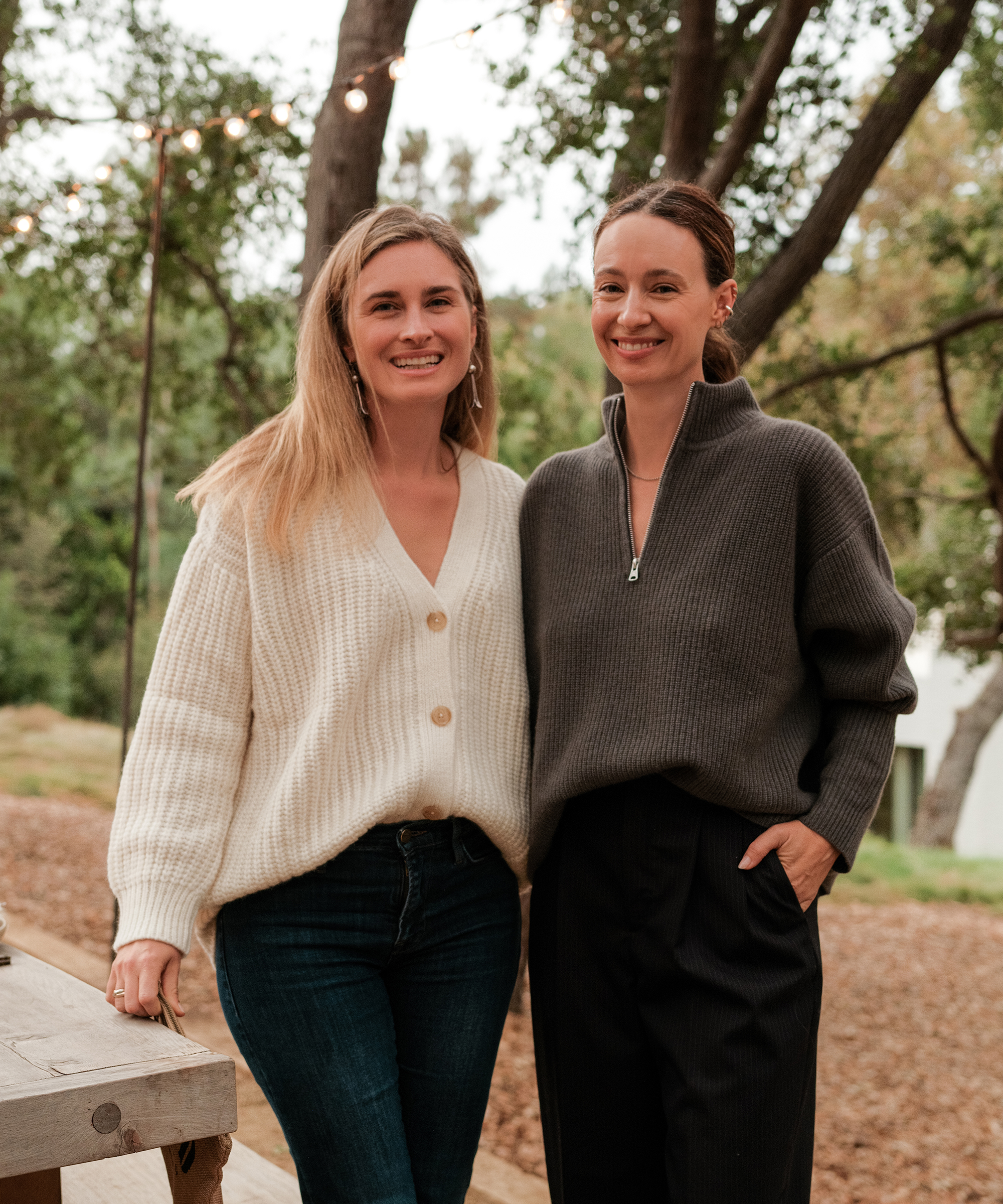Two women stand outdoors, smiling at the camera; one channels Lauren Bush Lauren’s casual style in a cream cardigan and jeans, while the other wears a gray zip-up sweater and black pants. String lights and trees glow softly in the background.