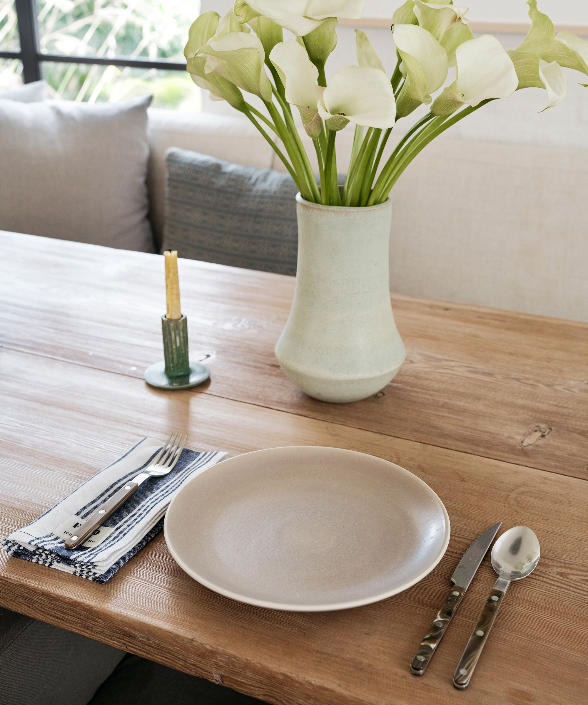 A wooden dining table set with a beige plate, silver cutlery on a striped napkin, a tall vase of white calla lilies, and a yellow candle in a green holder evokes the timeless style of Lauren Bush Lauren. Cushions are visible in the background.