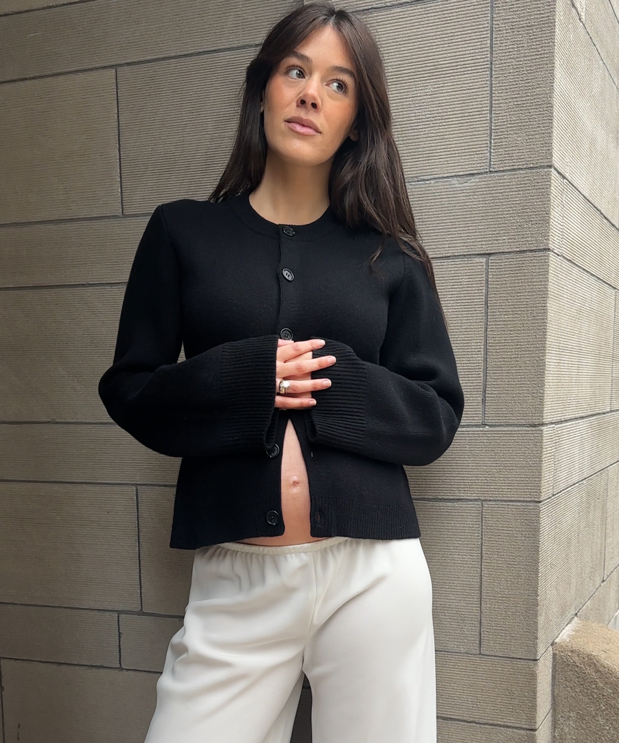 ilana in a black cardigan with white silk pants leaning against a stone wall outside
