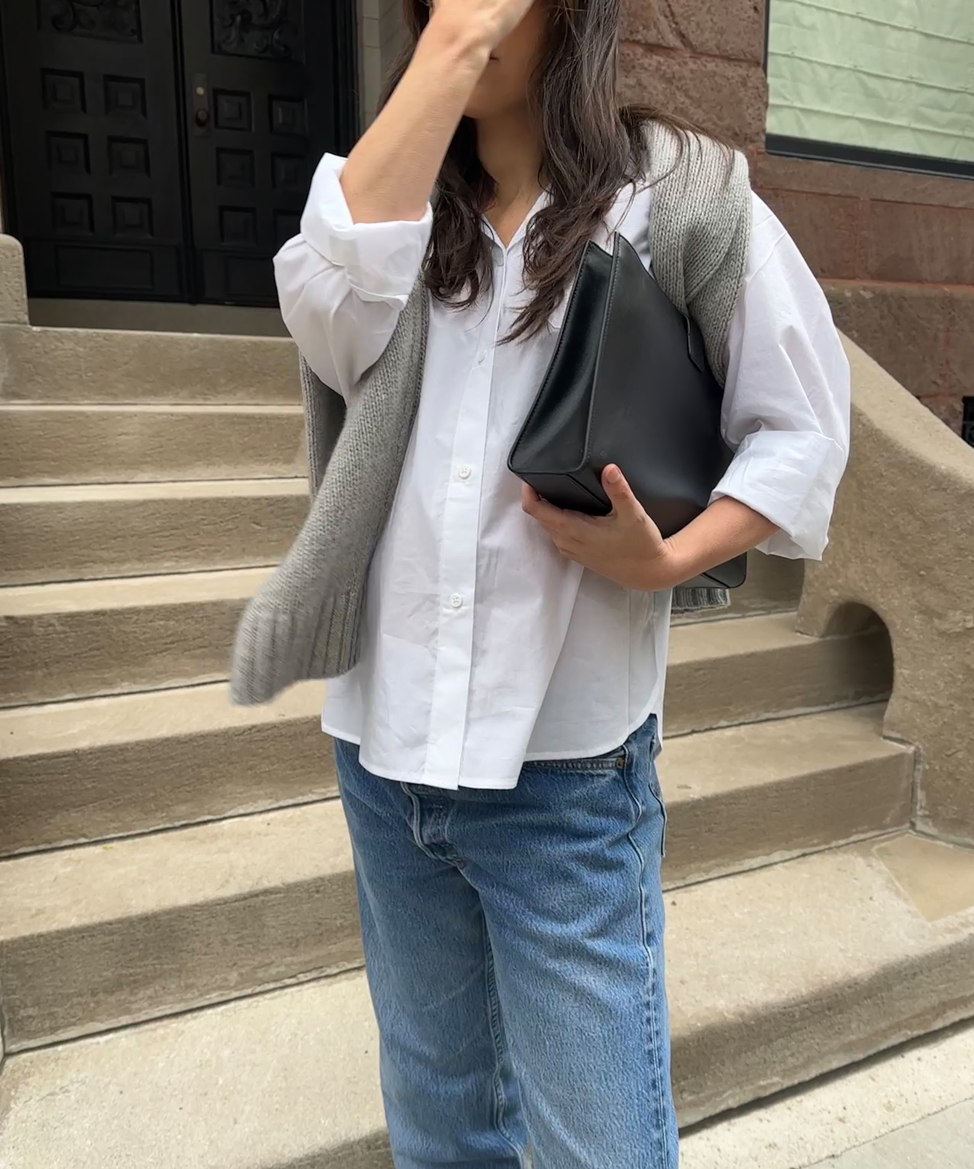 ilana in a white button up with blue jeans and a grey sweater over her shoulders holding a black leather bag standing on stone steps