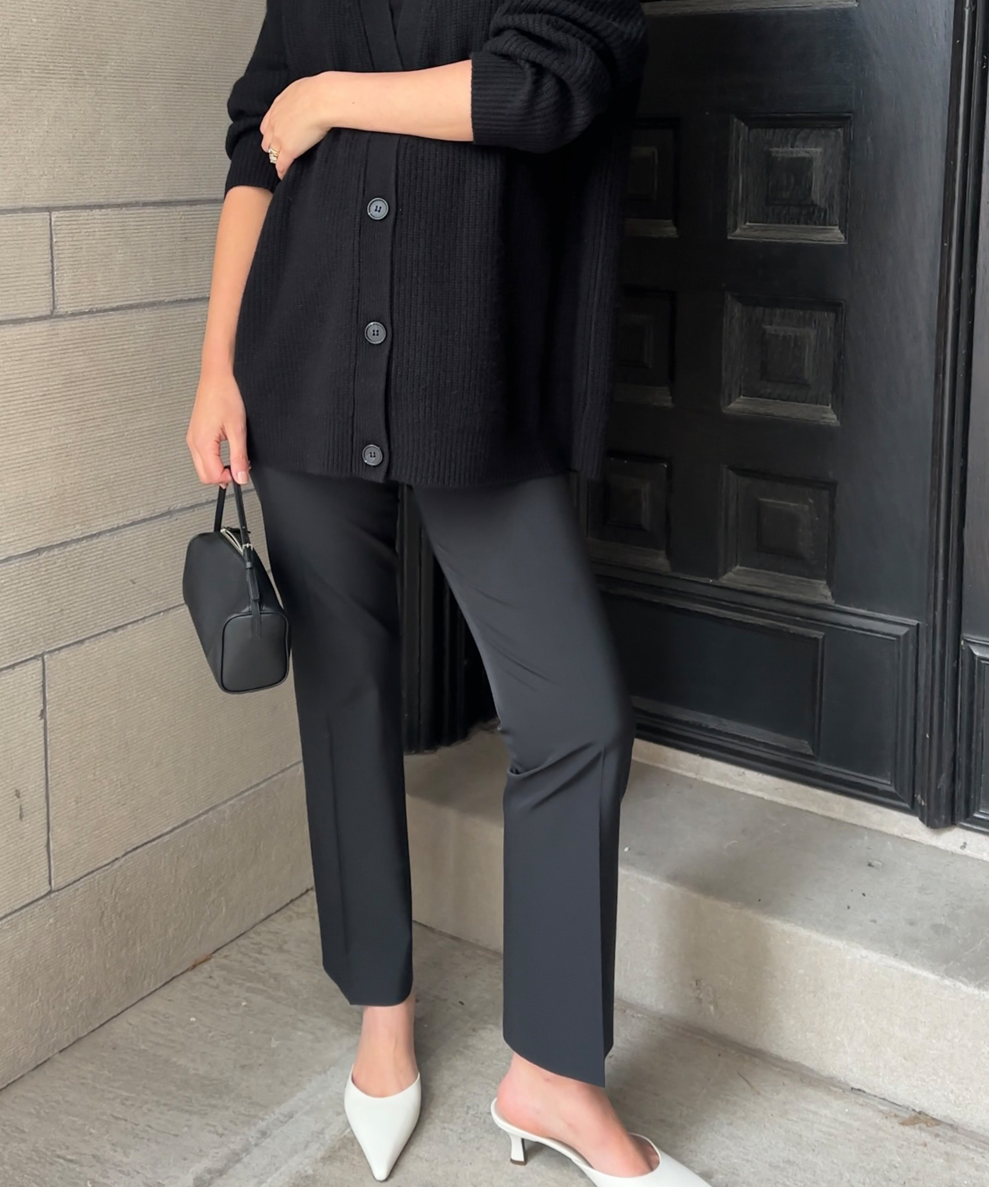 black cashmere cardigan with black trousers and white kitten heels standing in front of a black door