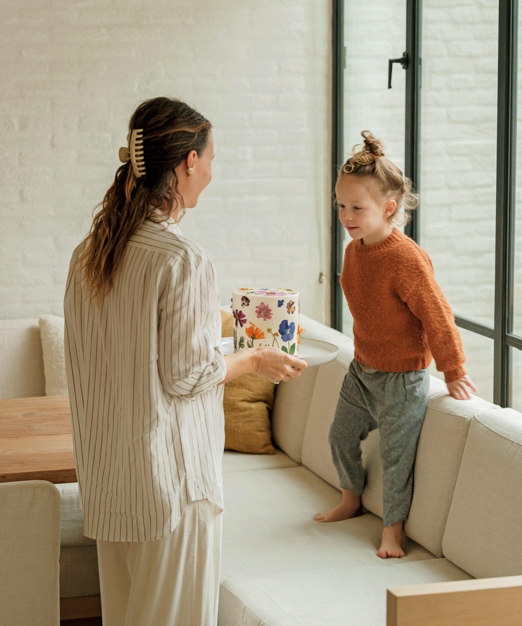 jenni in a striped boyfriend button up holding a cake in front of her son in her kitchen nook