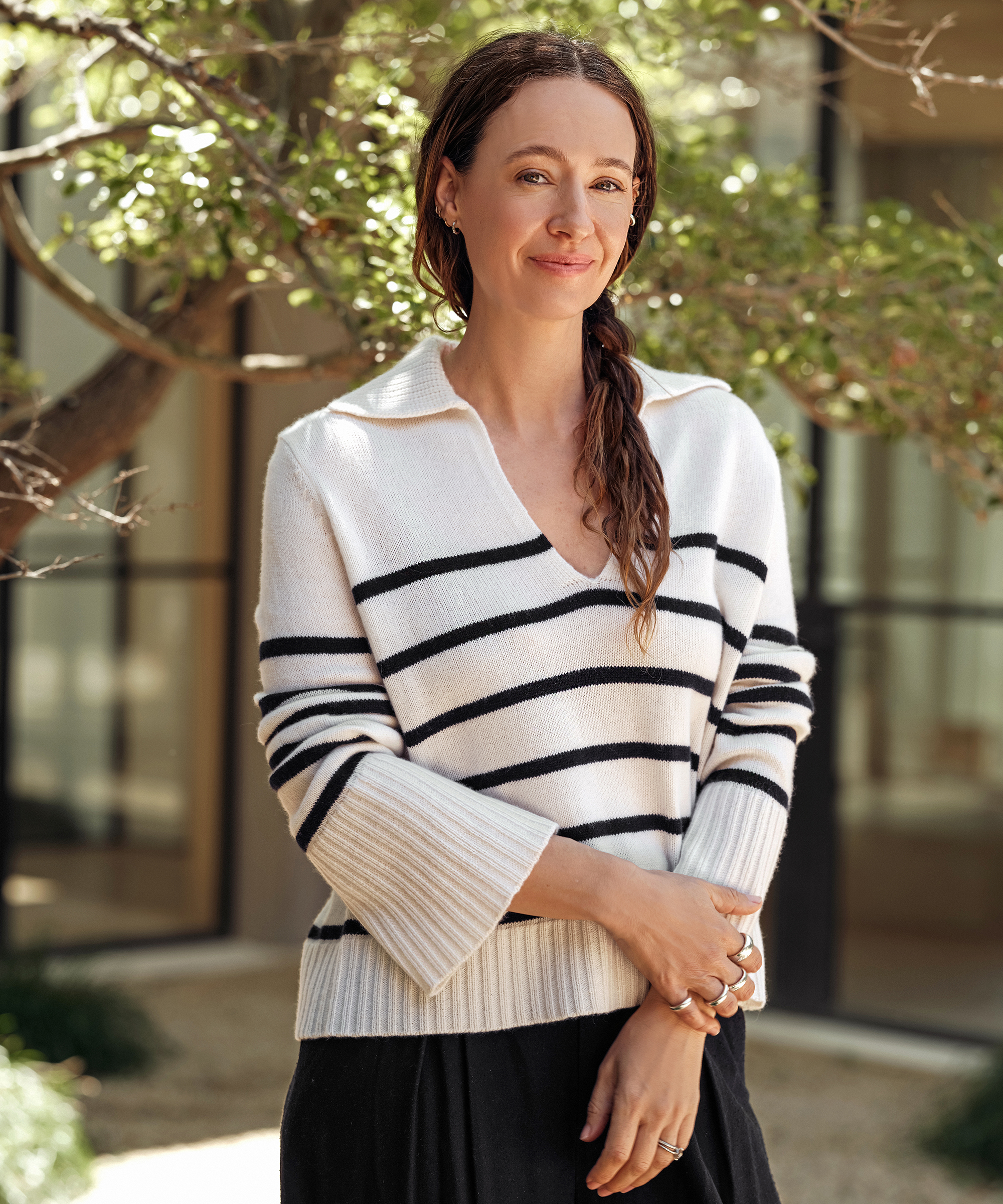 A woman with long brown hair stands outdoors, smiling slightly. She wears a summer outfit—a white sweater with black horizontal stripes and a black skirt. Green leafy branches and a building with large windows are in the background.