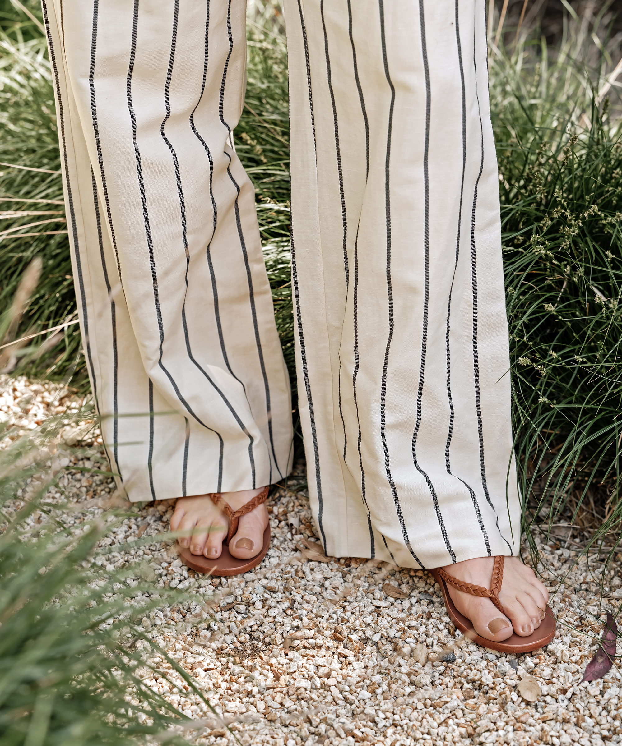 A person wearing a summer outfit of light-colored, wide-leg pants with vertical stripes and brown sandals stands on gravel among tall green grass.