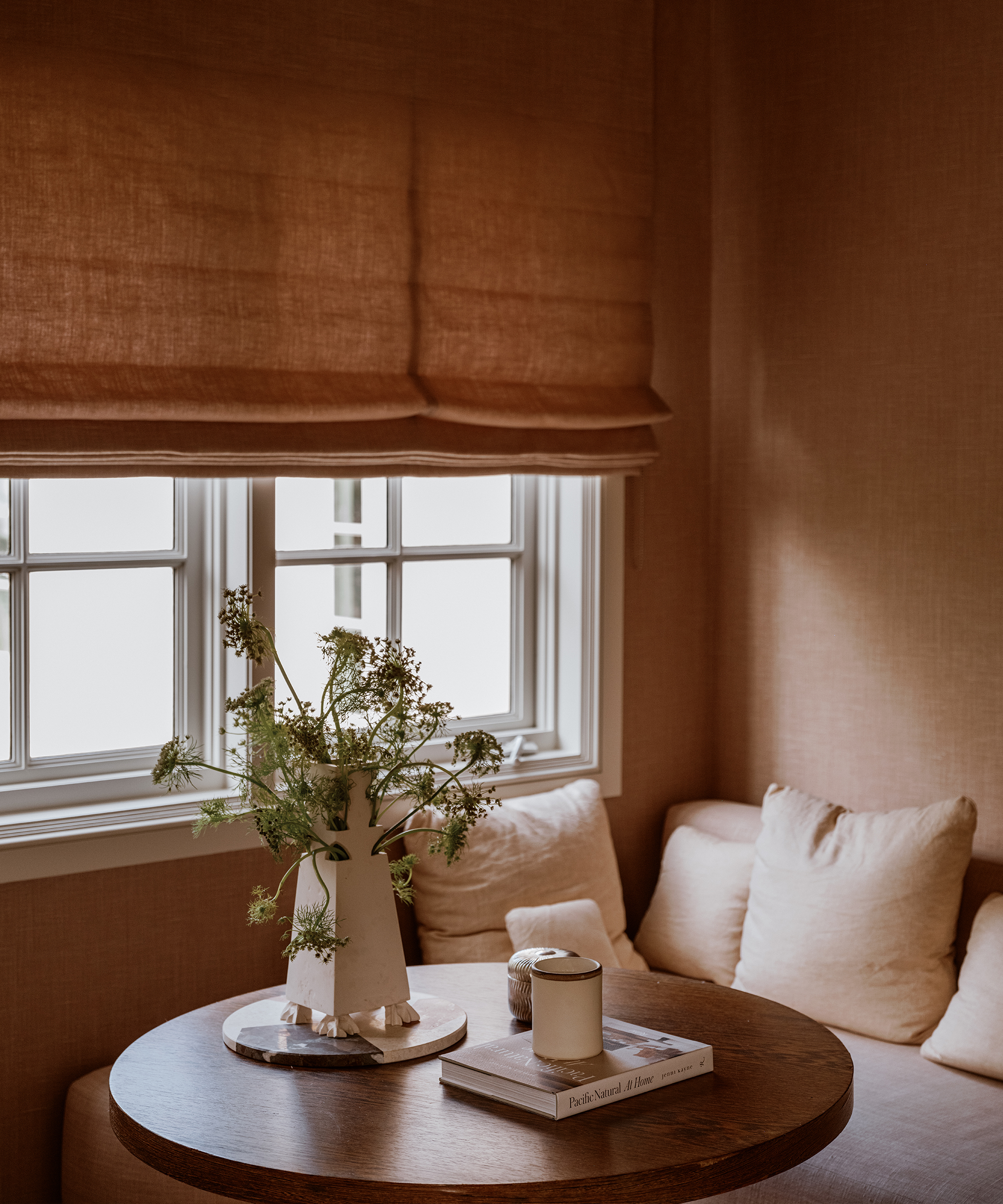 A cozy corner in Houston with a round wooden table holding a vase of greenery, a candle, and books. A cushioned bench with neutral pillows sits beside a window with brown Roman shades. Warm, soft lighting fills the space.