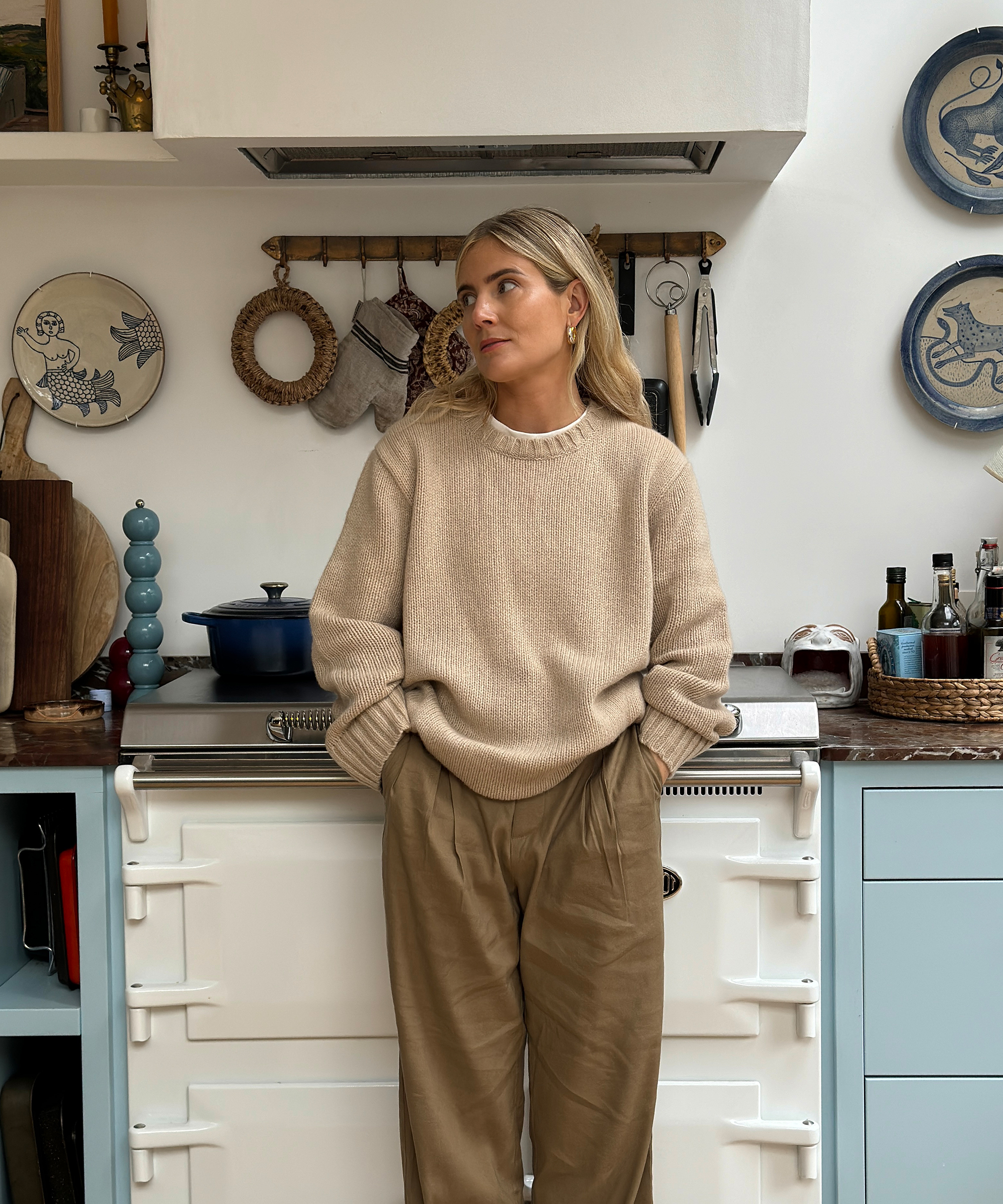 Lucy Williams, with blonde hair, stands in a cozy kitchen wearing a beige sweater and brown trousers. Decorative plates and cooking utensils hang on the wall behind her as she casually places her hands in her pockets.