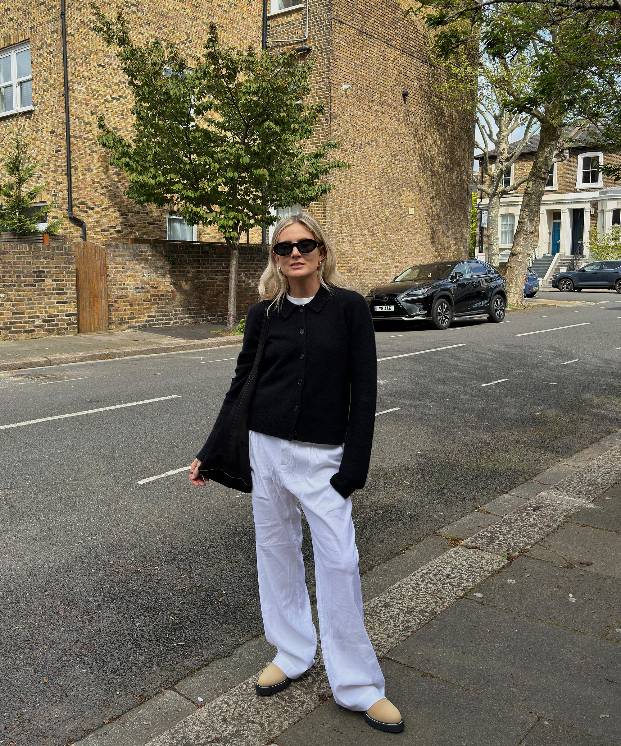 A woman inspired by Lucy Williams wears a black cardigan, white wide-leg pants, beige shoes, and sunglasses as she stands on a quiet residential street lined with brick buildings and parked cars.